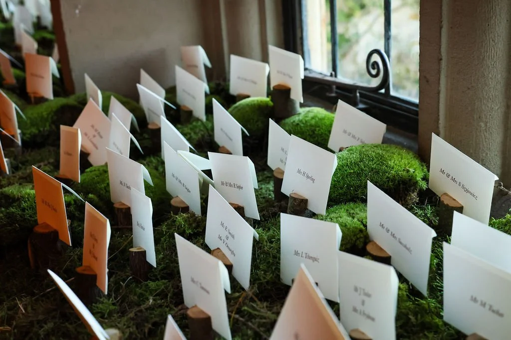 name and table cards with small white and orange place cards on them, arranged on moss-covered logs near a window.