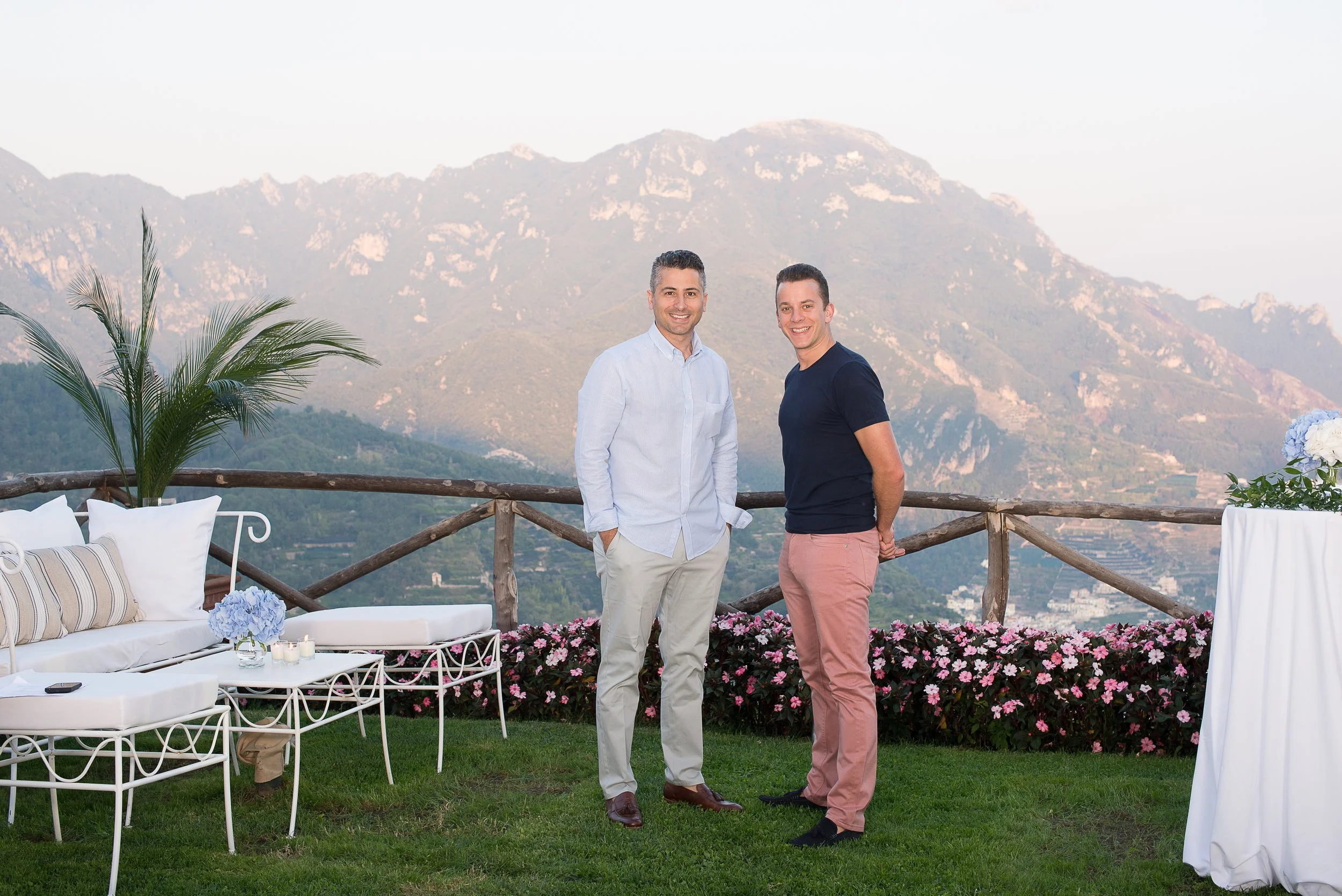 Just Seventy Stas Anastasiou and Laurie Weitzkorn standing outdoors on a grassy area with mountains in the background, surrounded by garden furniture and pink flowers, during a corporate cruise event