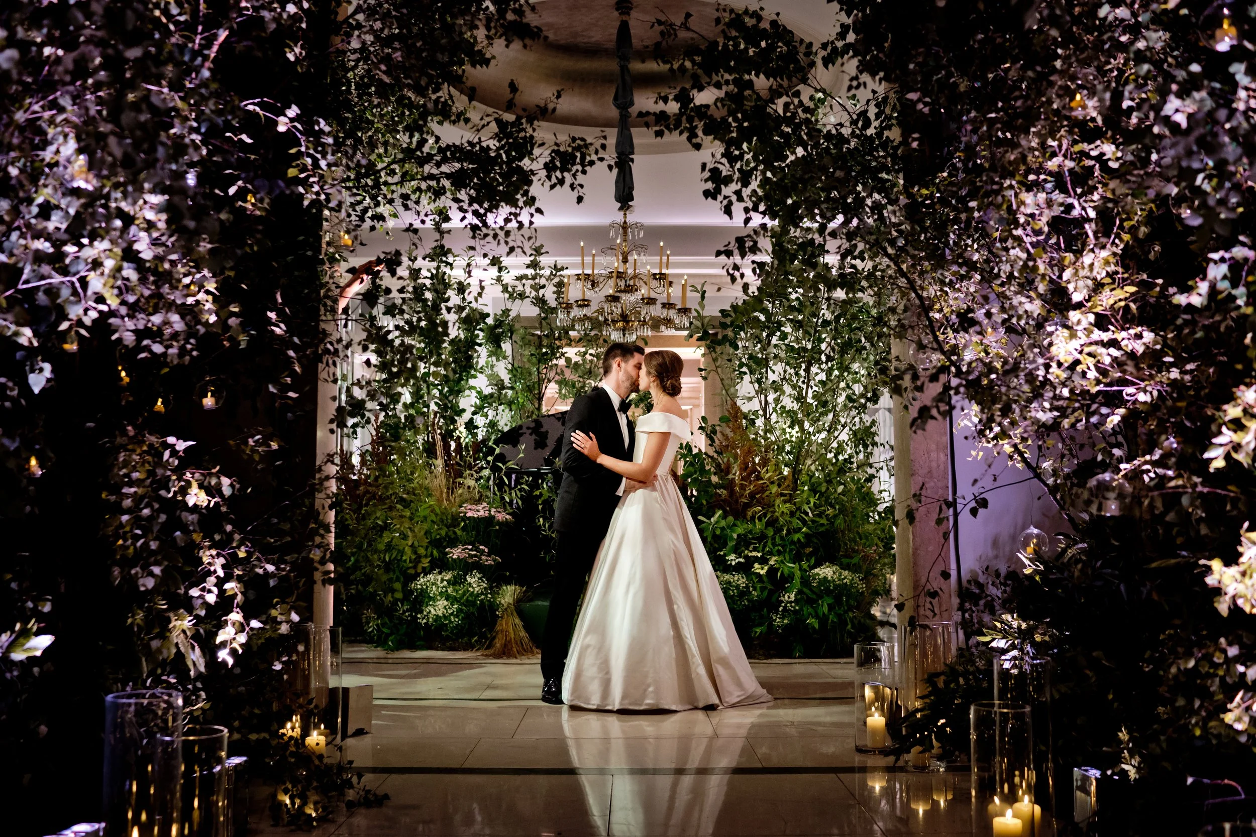 A bride and groom share a kiss during their wedding ceremony, framed by lush floral arrangements and illuminated by candles and a chandelier in an elegant indoor venue.