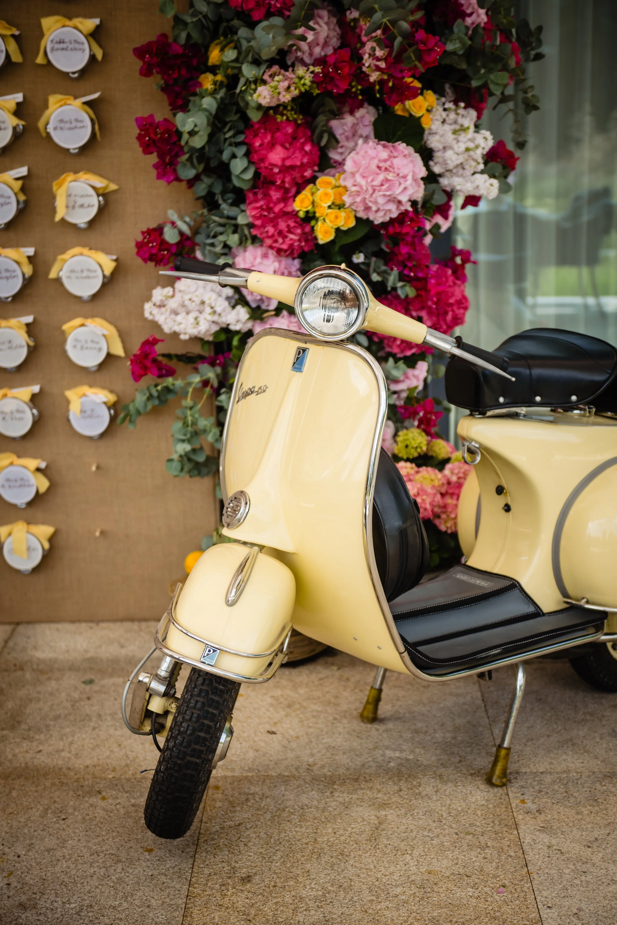 A vintage yellow Vespa scooter parked indoors near a floral display of pink, red, and yellow flowers. To the left, a board with small notes or cards attached with yellow ribbons. Fairmont Windsor Park'.