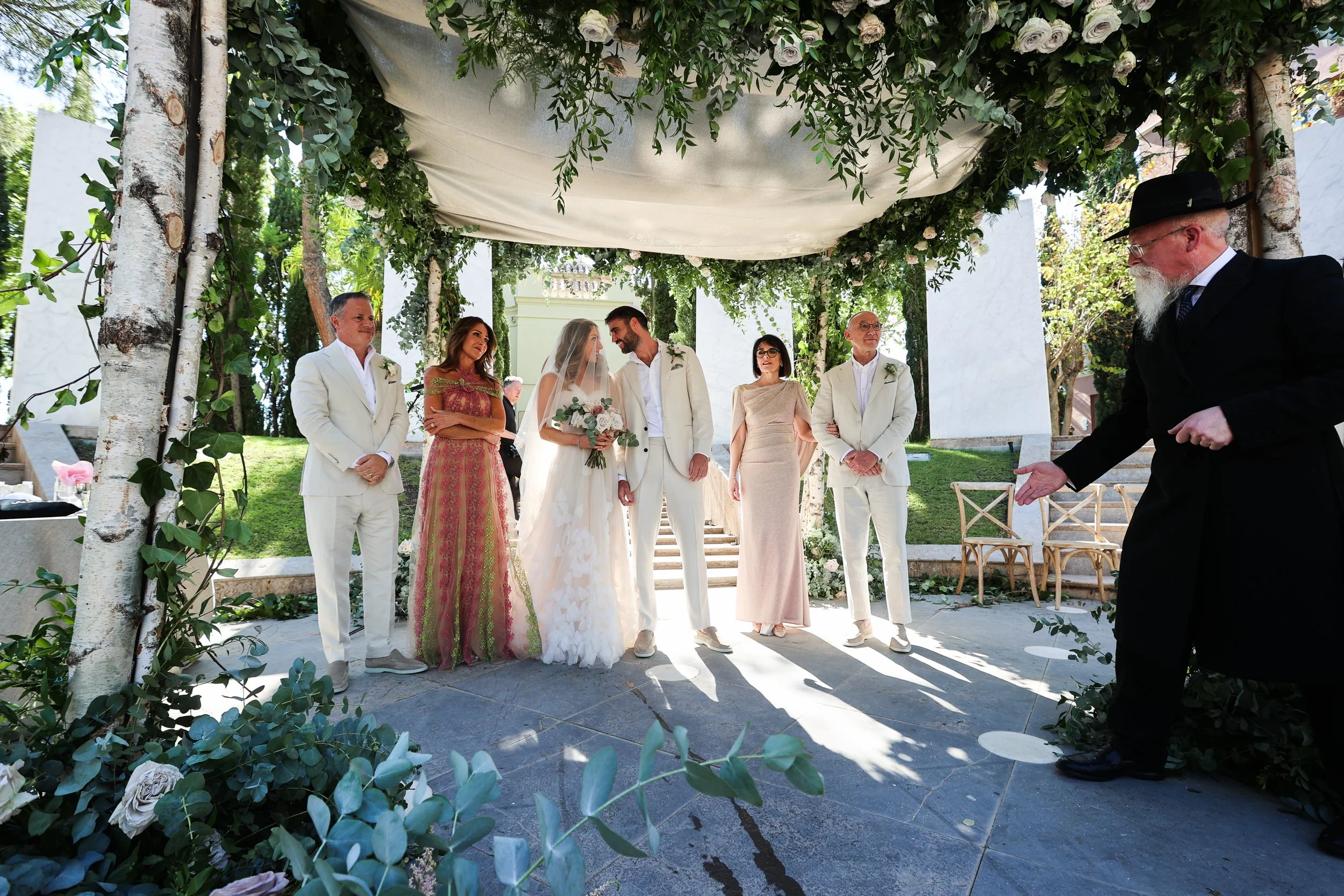 A wedding ceremony taking place outdoors with a bride and groom standing under a decorated chuppah. The scene is set on a grassy area with trees and modern white structures in the background at Anantara Villa Padierna Resort in Marbella