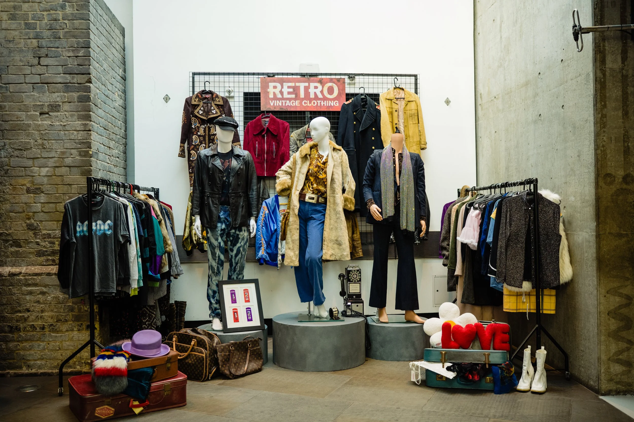 A vintage clothing store display with mannequins dressed in retro outfits, surrounded by racks of clothing, hats, and accessories, with a red sign that reads 'RETRO Vintage Clothing.'