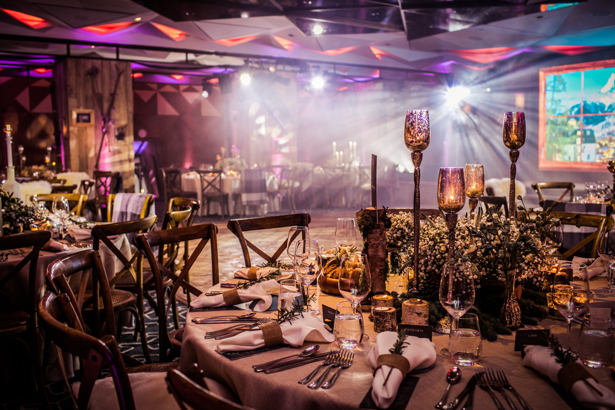 Elegant dining setup with round tables decorated with candles, floral arrangements, and glassware, set in a dimly lit banquet hall with colorful uplighting and a large window showing a scenic view.
