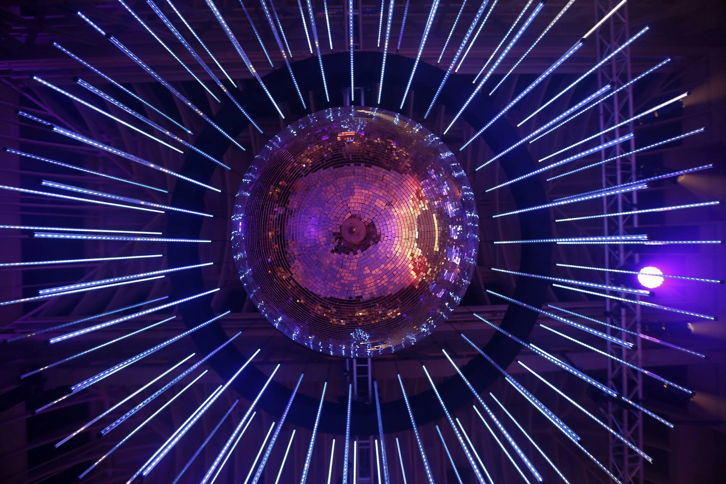 A large disco ball reflecting colorful lights, with blue LED light beams radiating outward in a dark environment.