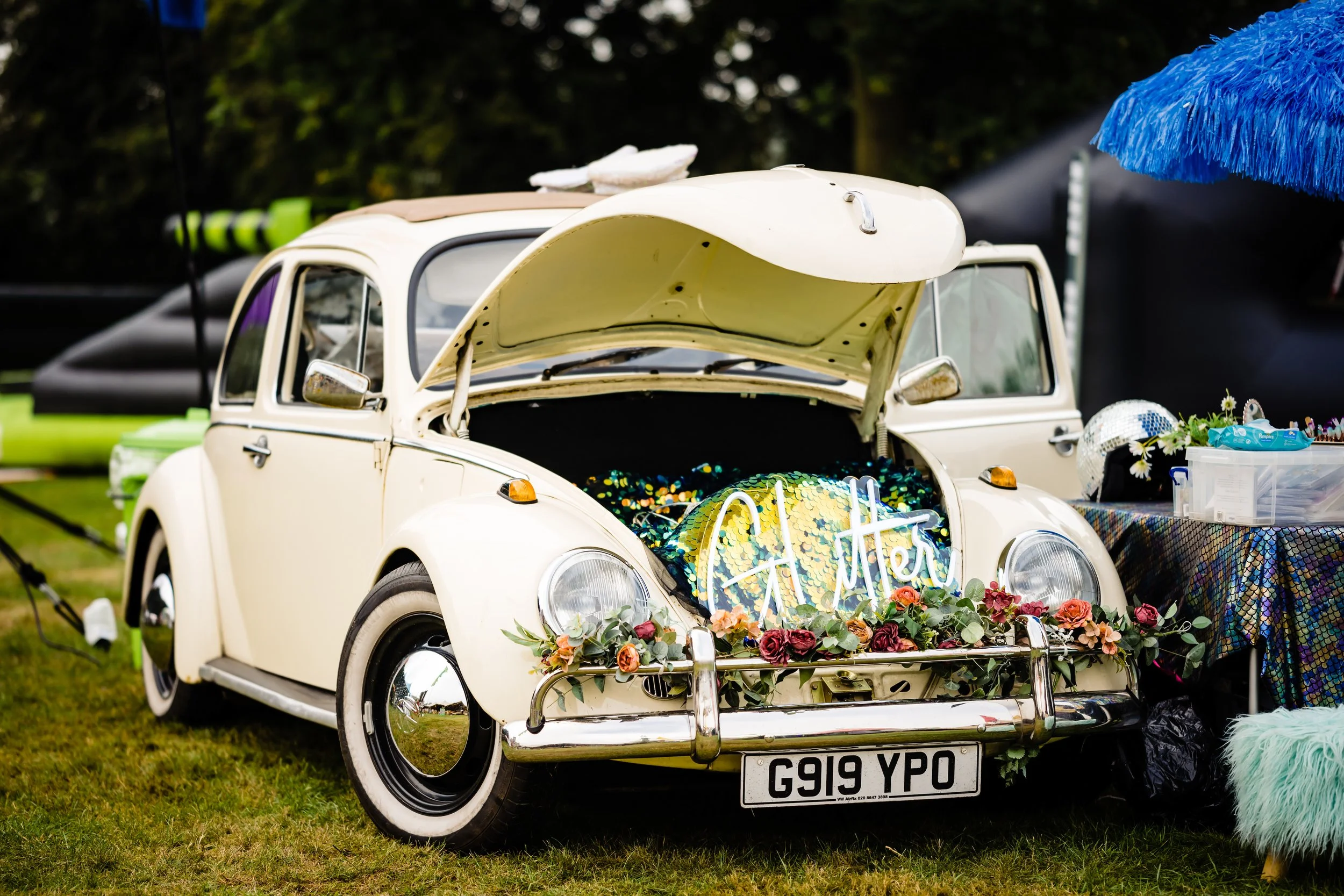 A white vintage Volkswagen Beetle decorated with flowers and a neon sign that reads 'Happily Ever After' on its open trunk, used as a display at an outdoor event at Bar Mitzvah at  Wrotham Park