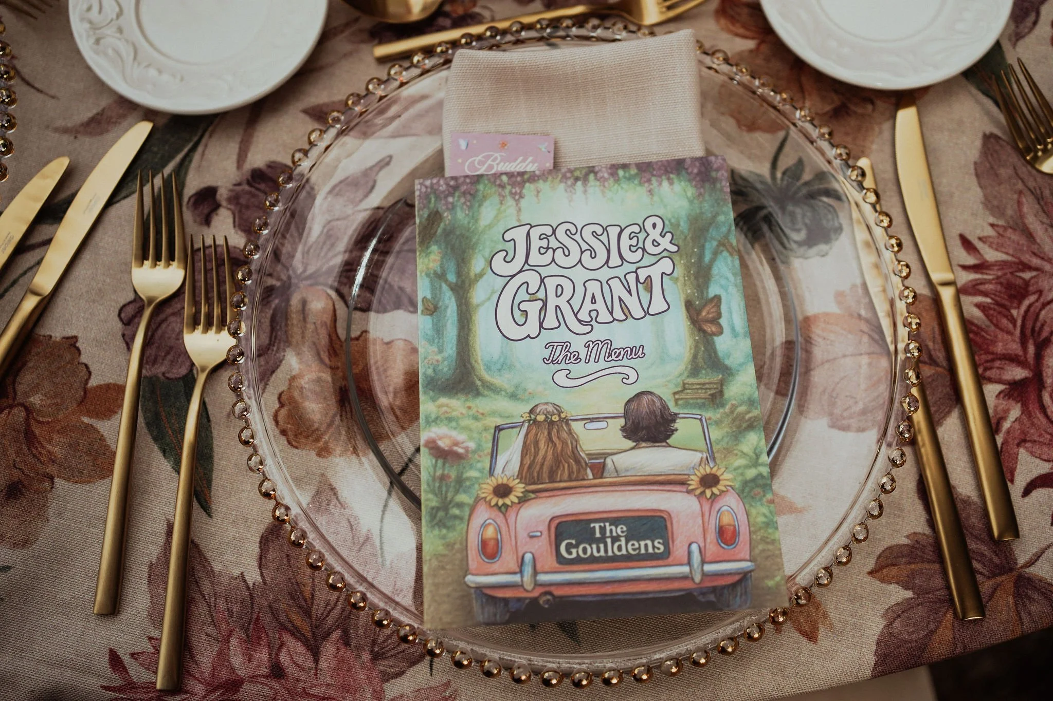 A clear glass plate with a beaded edge, set on a table with a floral tablecloth. There are gold-colored forks and knives on either side of the plate. On the plate, there is a folded beige napkin,  colorful booklet featuring an illustration of two peo
