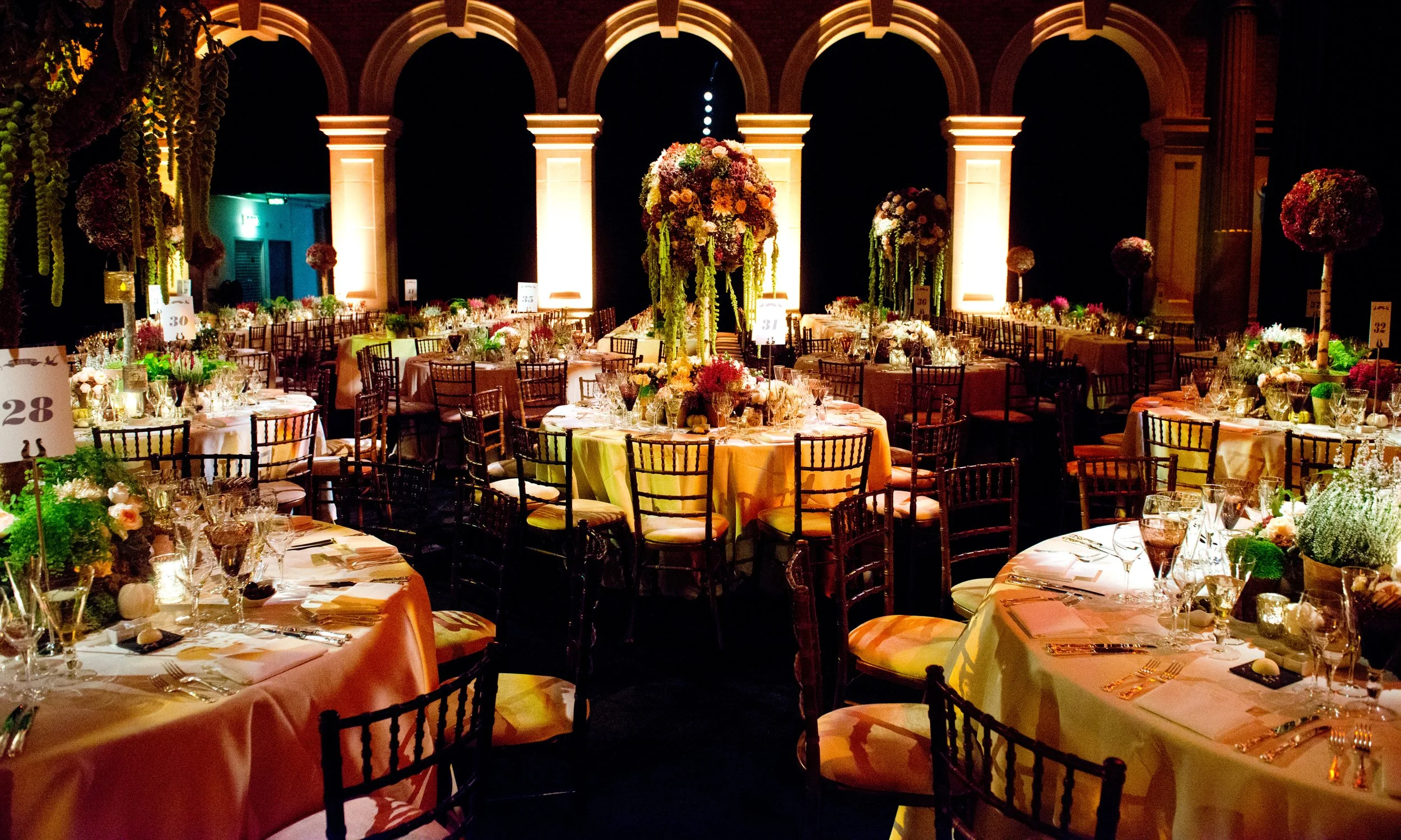 Elegant banquet hall decorated with floral centerpieces and set tables for a formal event, featuring high ceilings, arches, and warm lighting.