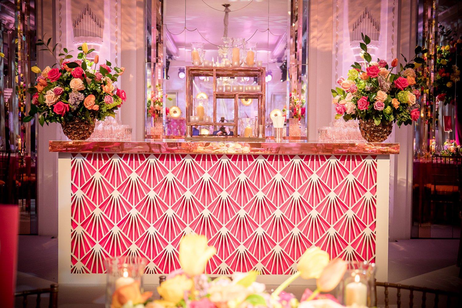 Decorative pink bar with floral arrangements, candles, and glassware in an elegant event setting.