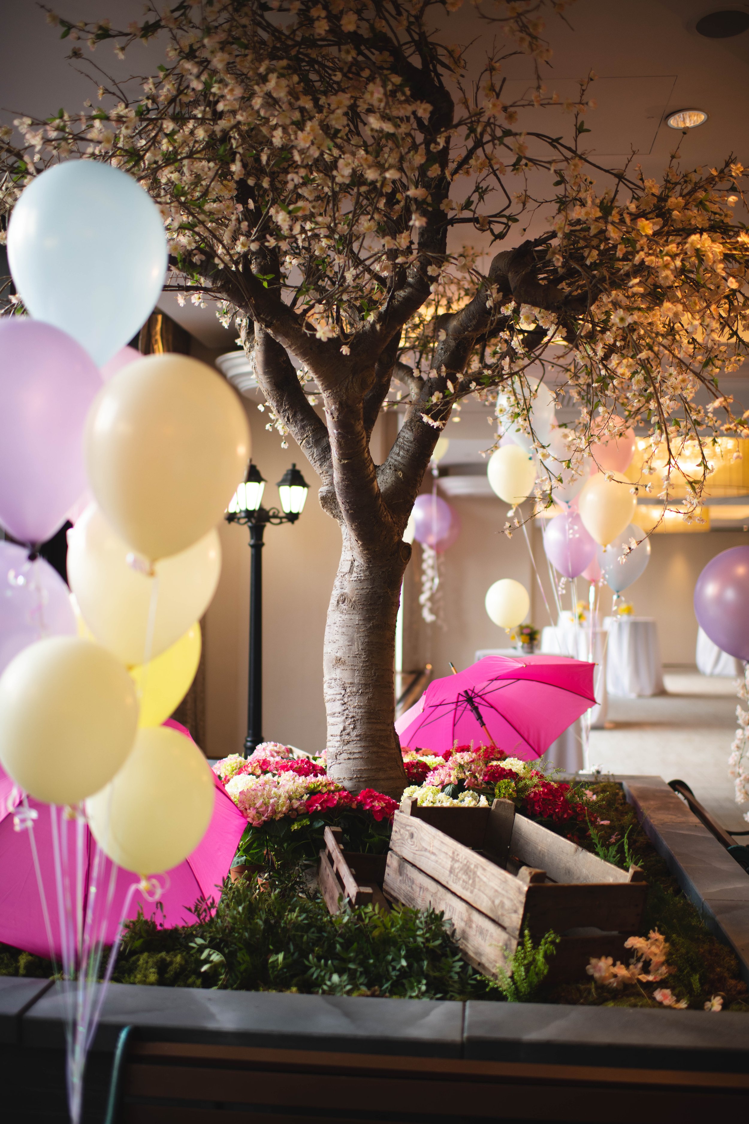 Indoor scene featuring a central artificial tree with blooming pink and white flowers, surrounded by pastel-colored balloons, a pink umbrella, and flower arrangements in a wooden planter box. Soft lighting and decorative lamps create a festive atmosp