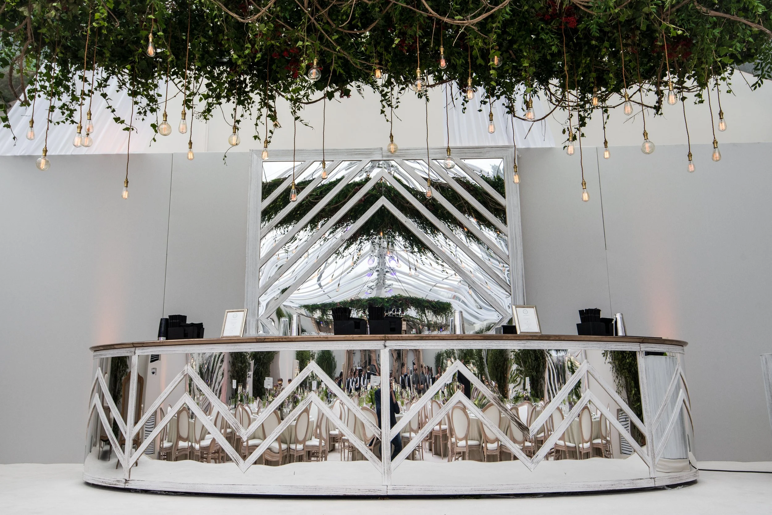 Interior view of a modern space with hanging light bulbs, a large mirror reflecting a decorated ceiling with greenery, and a decorative white wooden fence in the foreground.