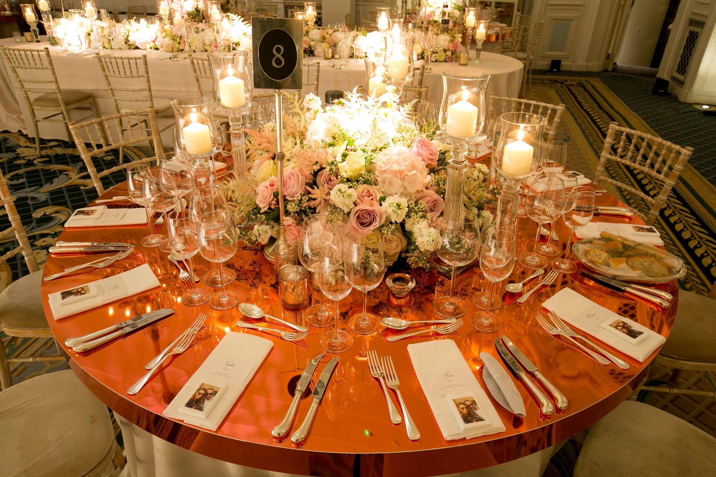 A round banquet table decorated with a large floral centerpiece of pink and white flowers, surrounded by lit candles in glass holders, set with wine glasses, silverware, and white napkins, in an elegant event hall.