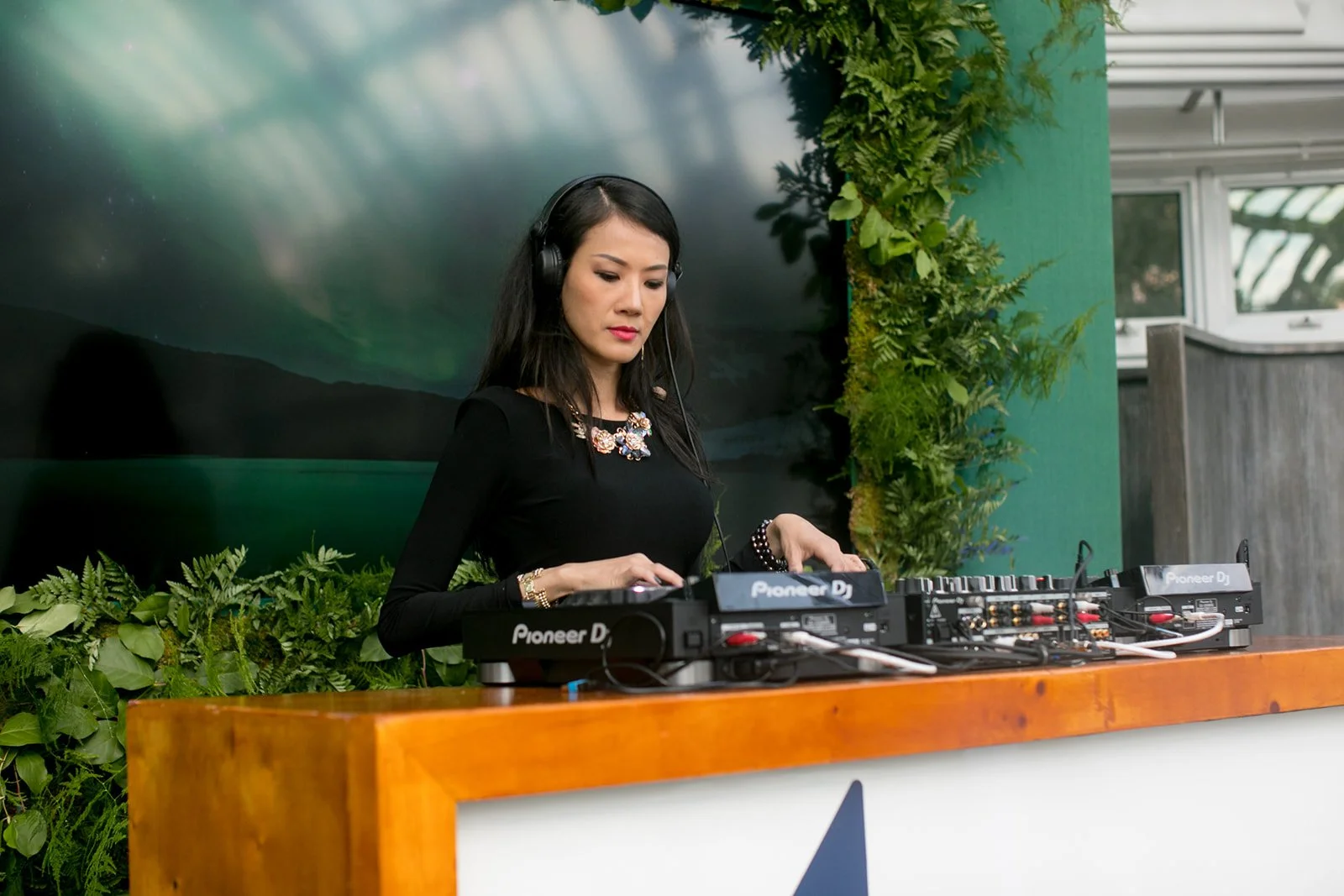 A woman DJ wearing headphones and a black dress mixing music at an outdoor event, with green plants and a black screen background.