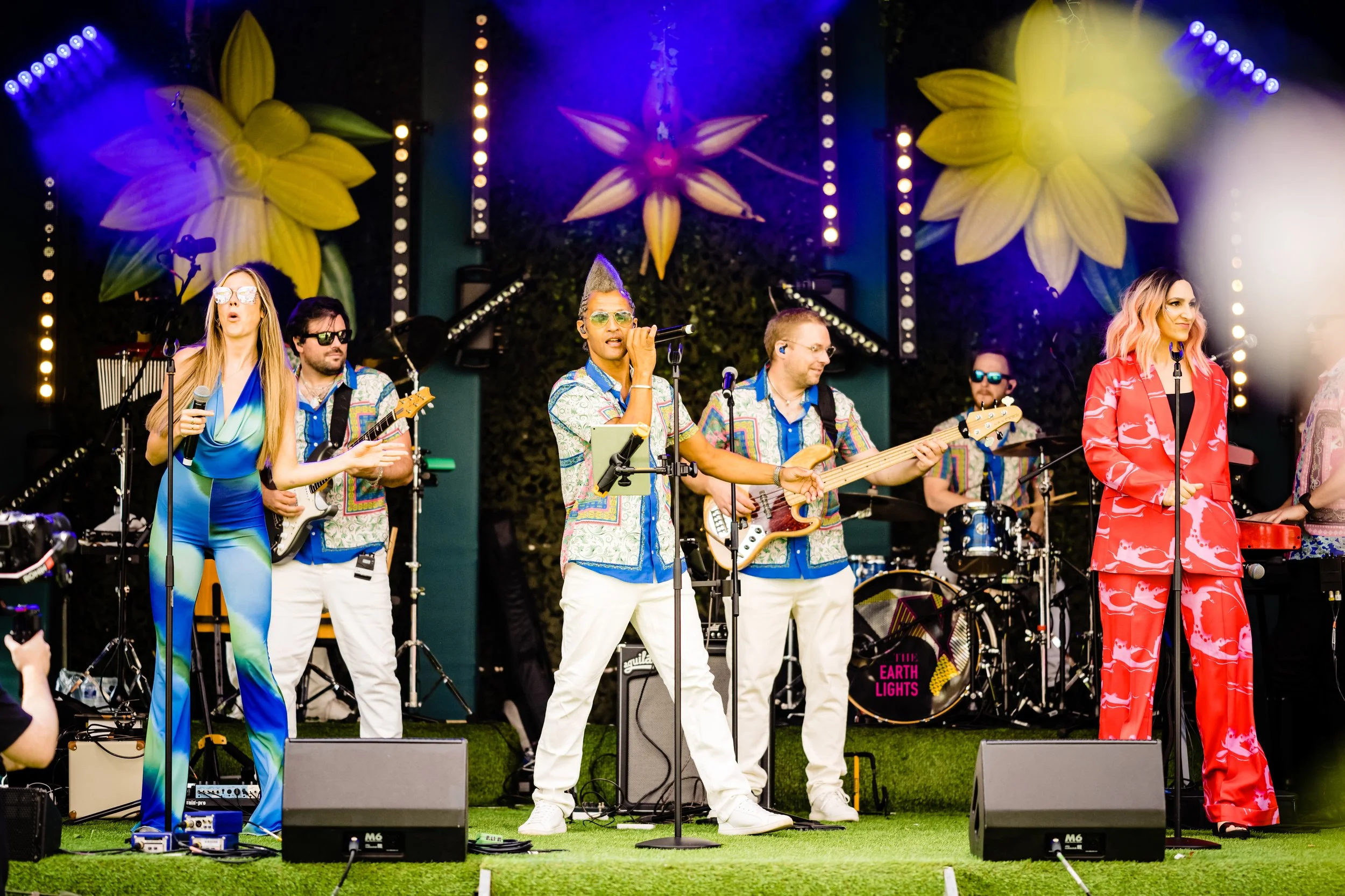 A band performing on stage with large yellow flower decorations in the background, colorful lights, and members wearing vibrant clothing at a Bar Mitzvah at  Wrotham Park