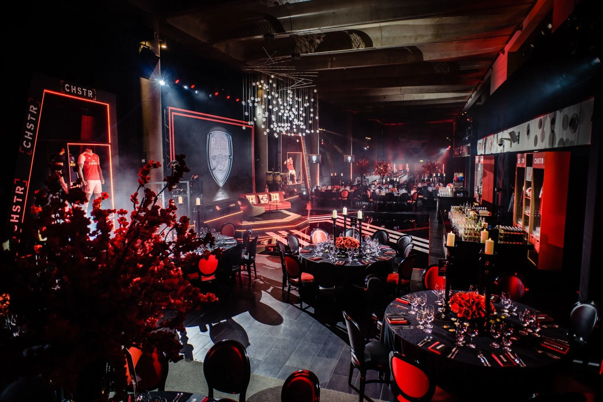 A decorated event space with black and red color theme, round tables with black tablecloths, red chairs, floral centerpieces, candles, and a stage with a large screen displaying an Arsenal logo, chandeliers, and lighting effects.