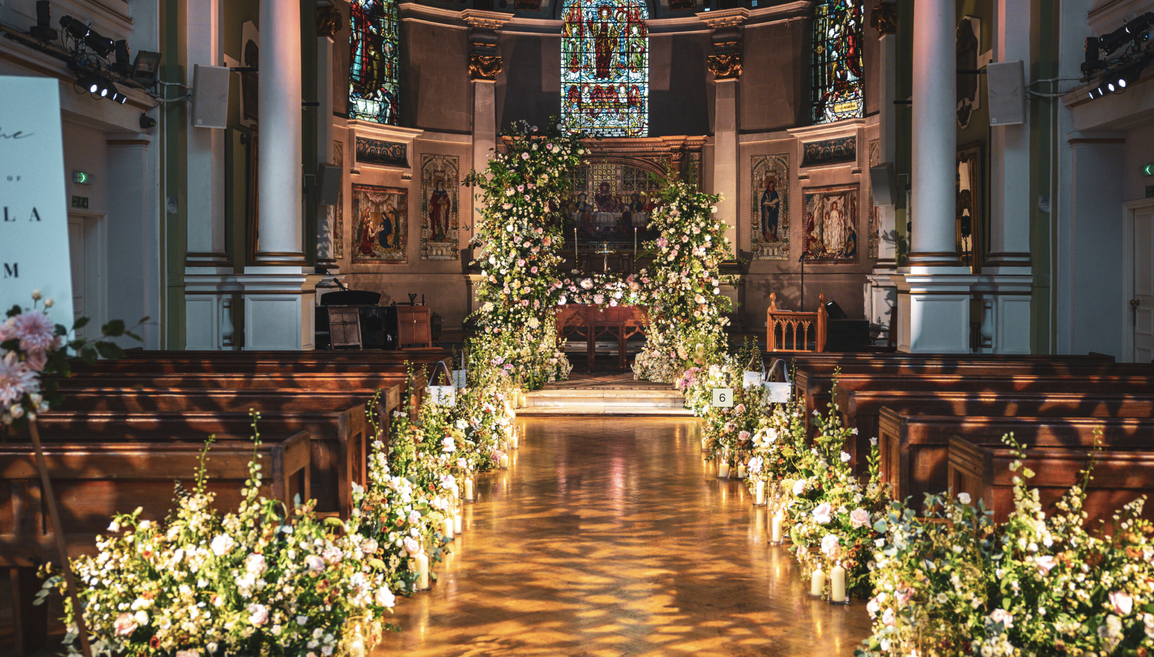One Marylebone church decorated for a jewish wedding with floral arch, pews lined with flowers, stained glass windows, and candle decorations along the aisle.