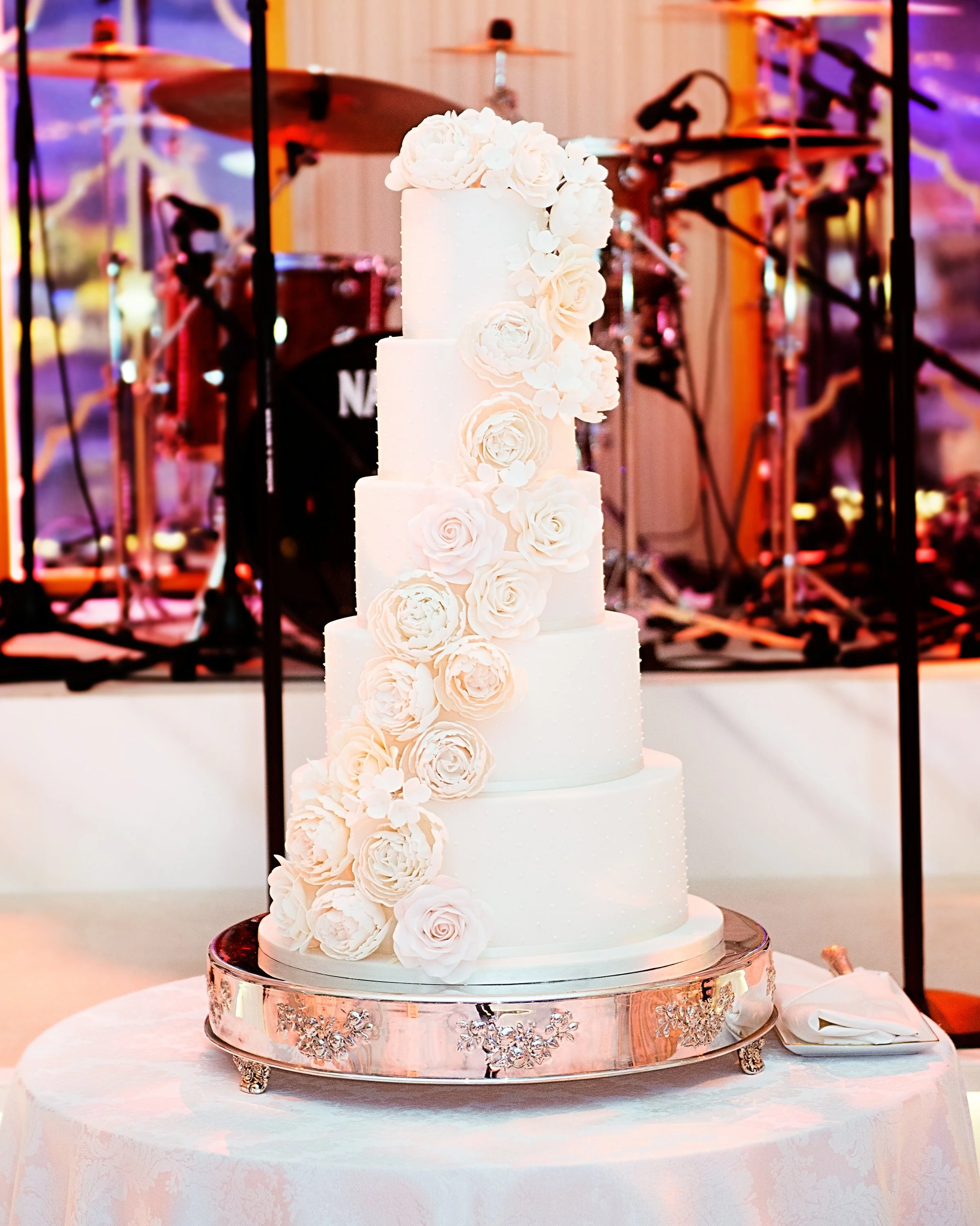 A tall, white, multi-tiered wedding cake decorated with white roses and flowers, placed on a silver cake stand on a round table with a white tablecloth. In the background, there is a drum set and musical equipment.