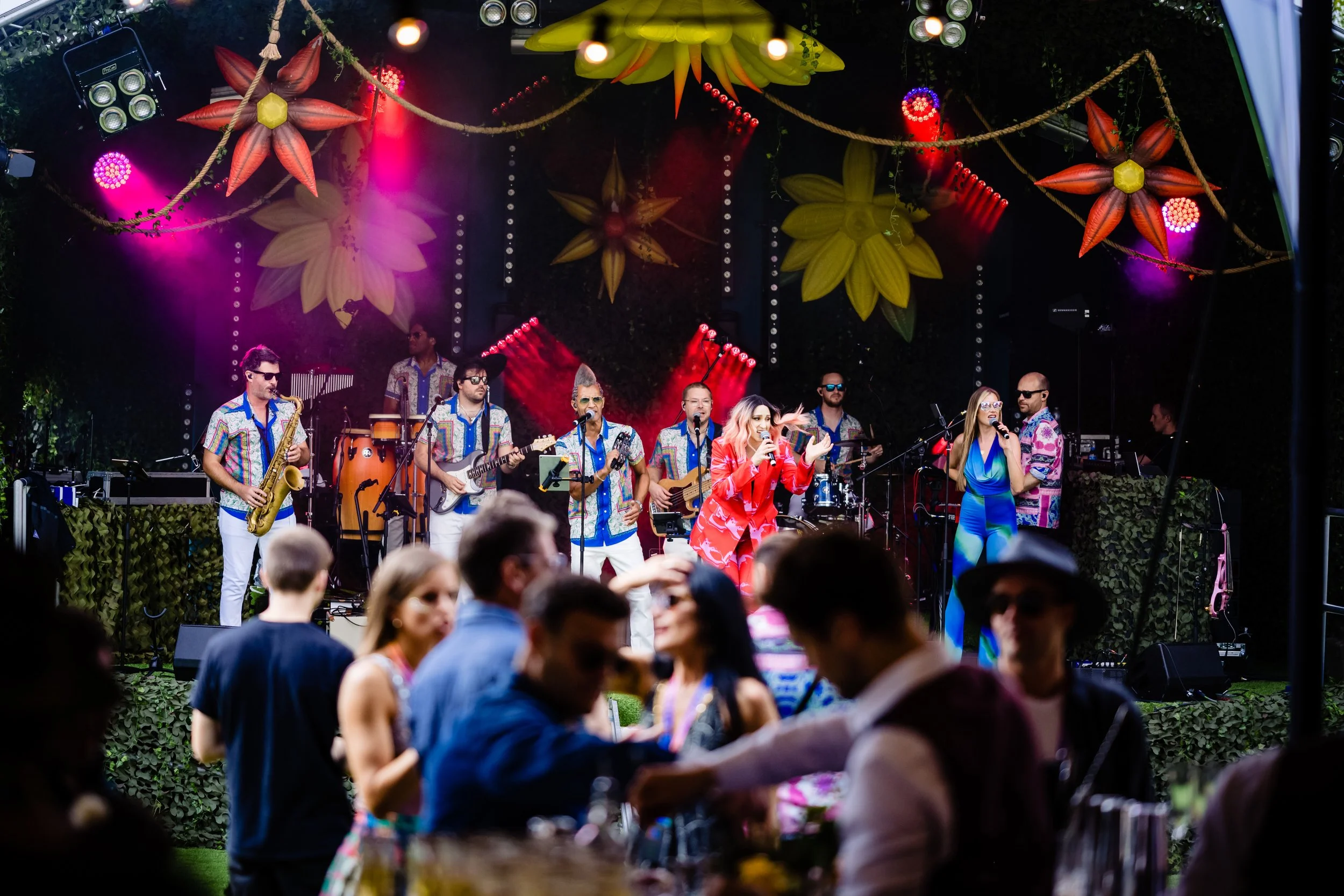 A band performing live on an outdoor stage decorated with large colorful flowers and hanging lights, with an audience enjoying the show.