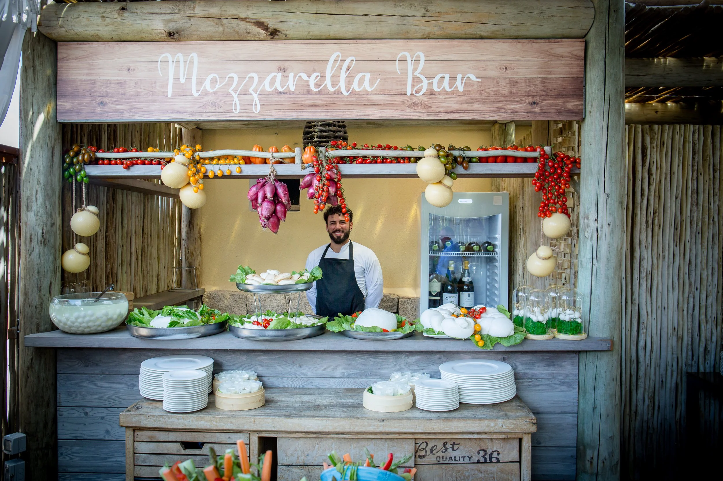 A man smiling at a mozzarella cheese stand decorated with cherry tomatoes, garlic, and other vegetables. The stand has a wooden sign reading 'Mozzarella Bar' and various cheese and salad items displayed.