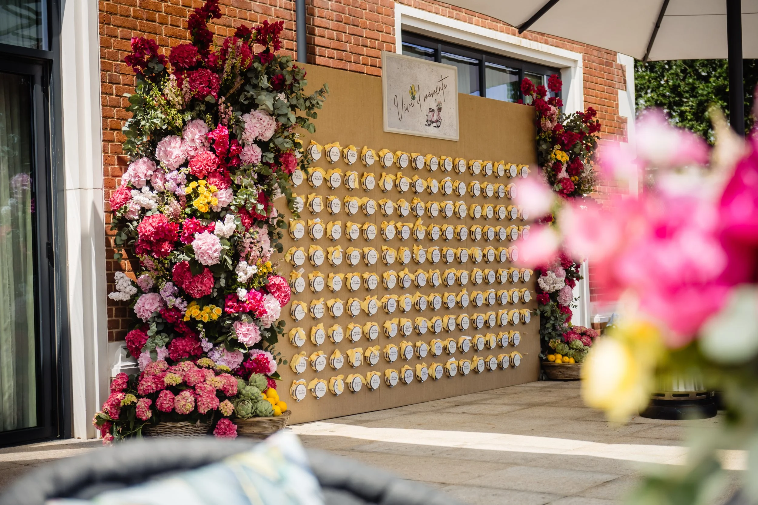 A decorative wall with multiple small yellow pouches and a sign that reads 'Vivi il momento' surrounded by vibrant pink and red flowers, with a brick building in the background. Fairmont Windsor Park'.