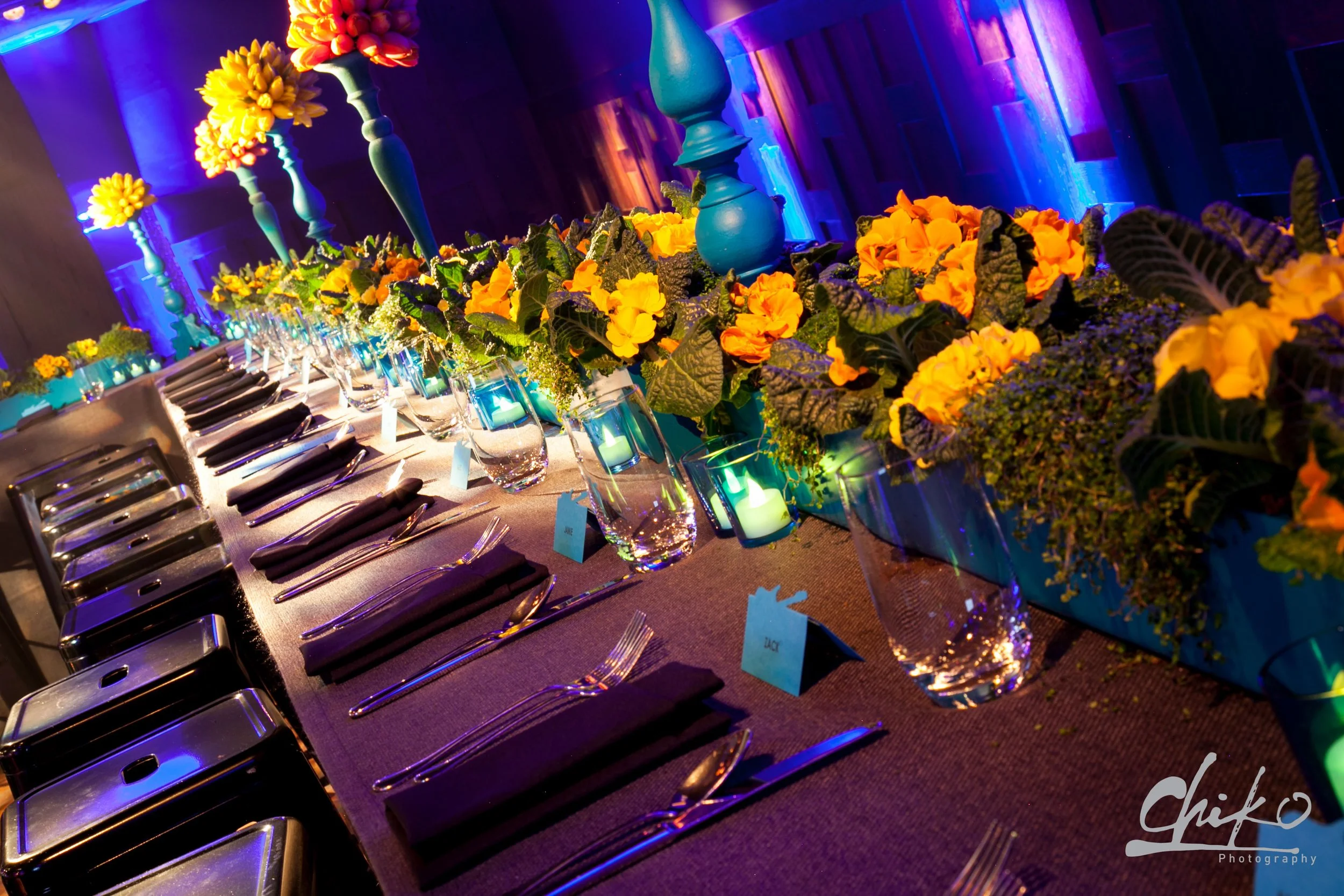 A beautifully decorated banquet table with a floral centerpiece featuring yellow and orange flowers, surrounded by blue candles and elegant tableware in a dimly lit room with vibrant multicolored lighting.