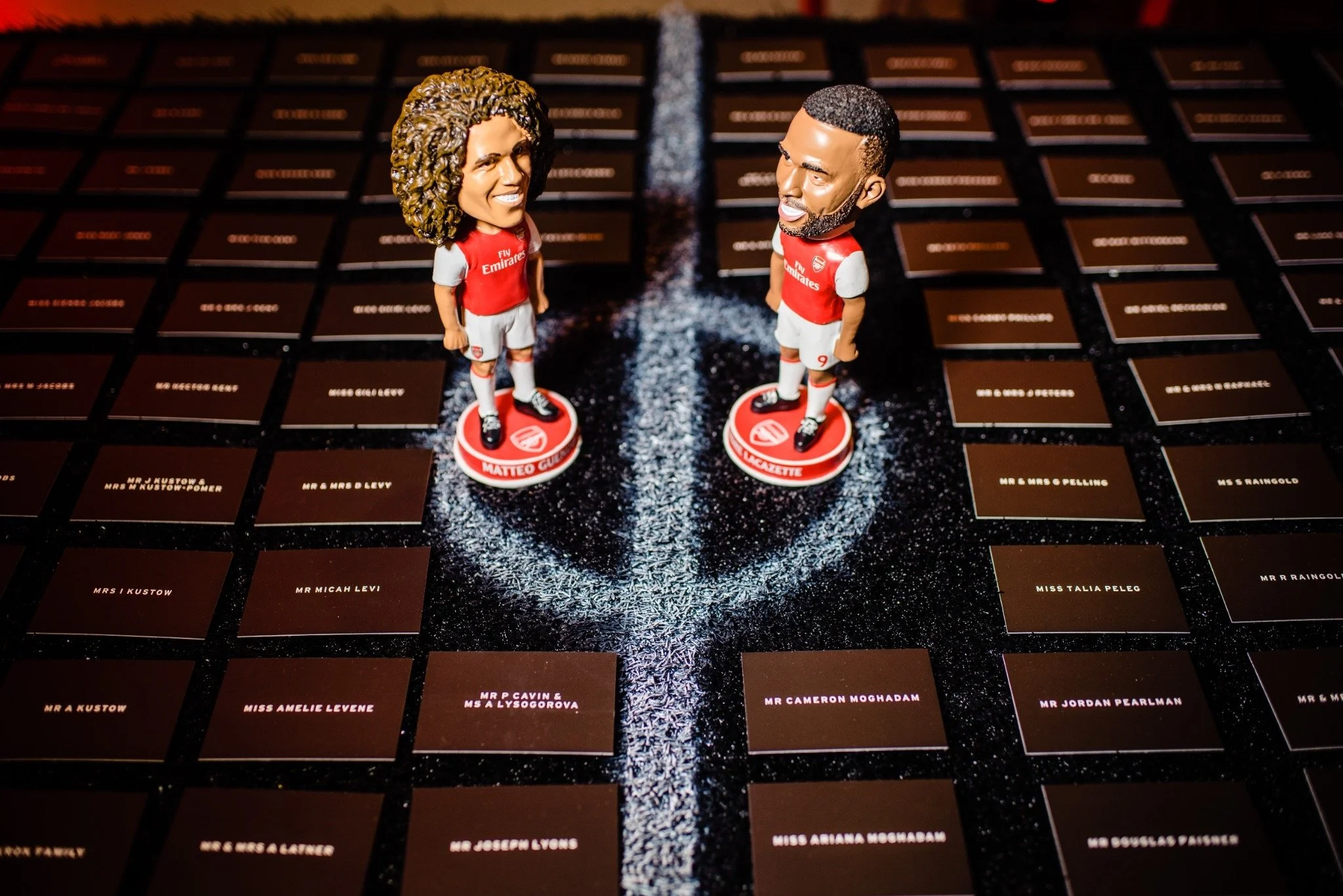 Two figurines with large heads, wearing red soccer jerseys and white shorts, standing on a black and brown background with name tags.