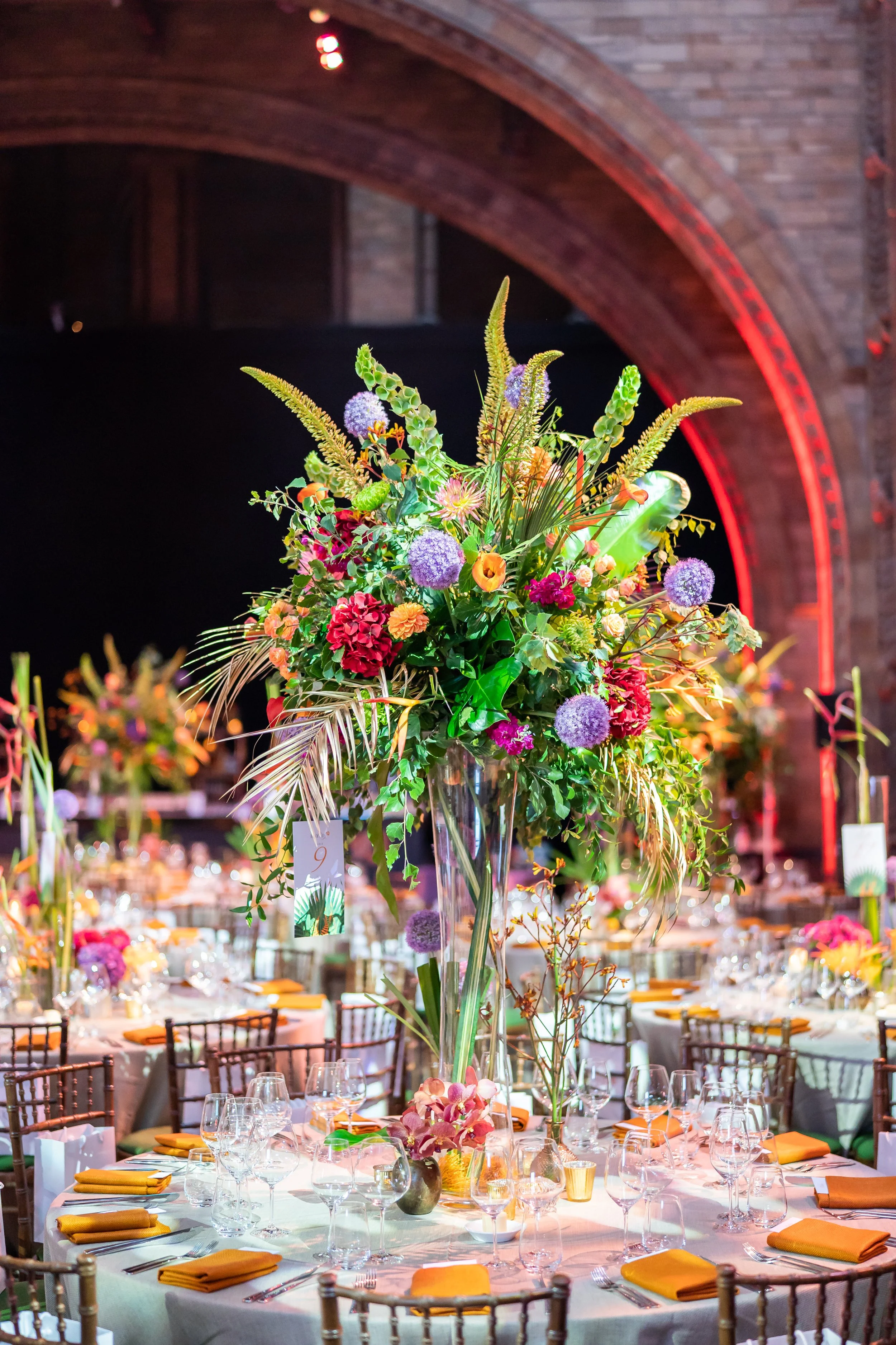 A floral arrangement on a tall glass vase as part of a corporate awards table setting. The arrangement includes purple, pink, orange, and green flowers with greenery.Background features a brick wall and arched ceiling at The natural History Museum