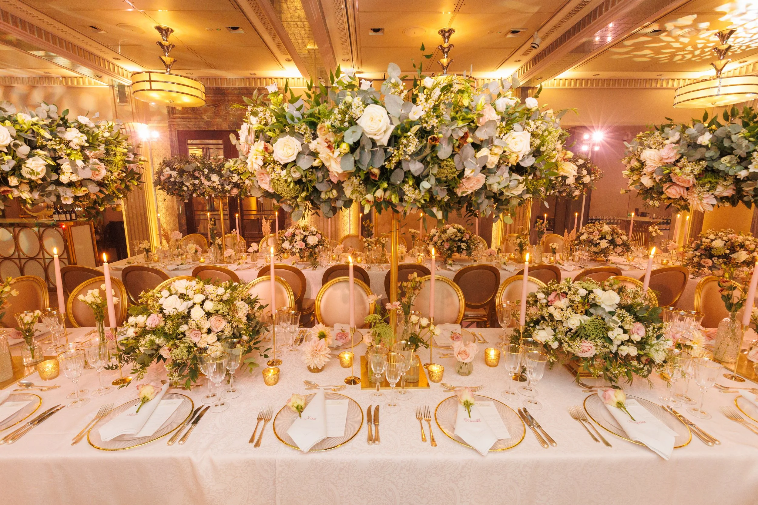 Elegant banquet table decorated with large floral arrangements of white and soft pink flowers, greenery, and candles in small holders in a decorated event hall