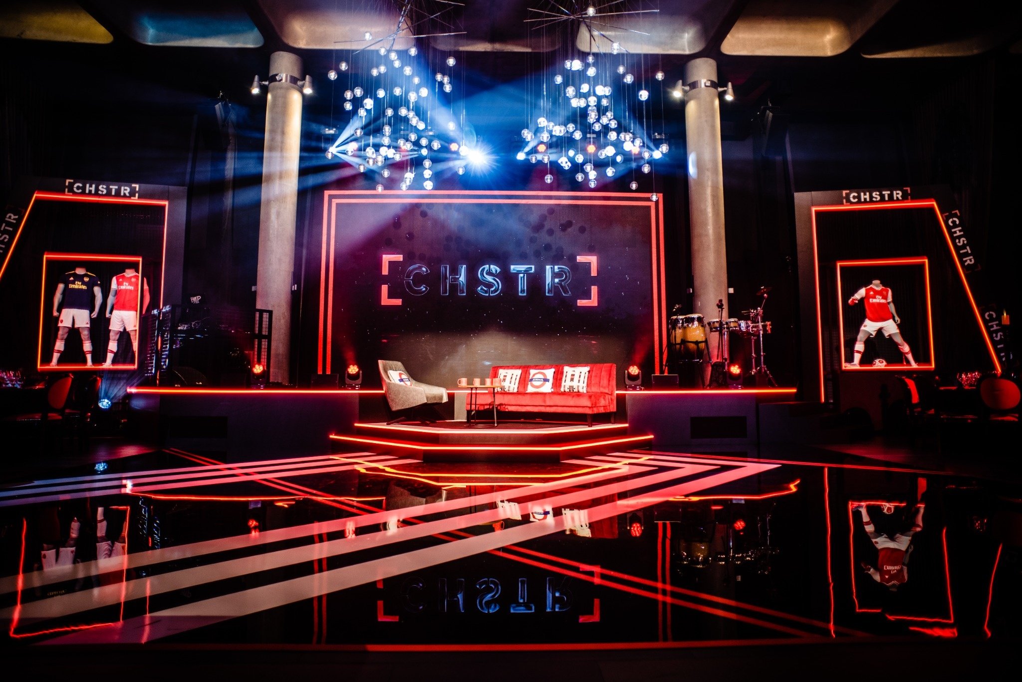 A stage with illuminated red outlines, sports jerseys displayed on mannequins, and a large digital screen showing the word "CHSTR". A modern chandelier with hanging glass globes is above, and musical instruments are on the right. The scene is reflect