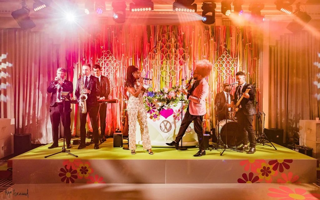 A lively band performs on a decorated 70s stage with colorful floral decorations, pink and orange curtains, and stage lighting. Musicians include a saxophonist, keyboardist, guitarist, drummer, and two women singing or speaking into microphones.