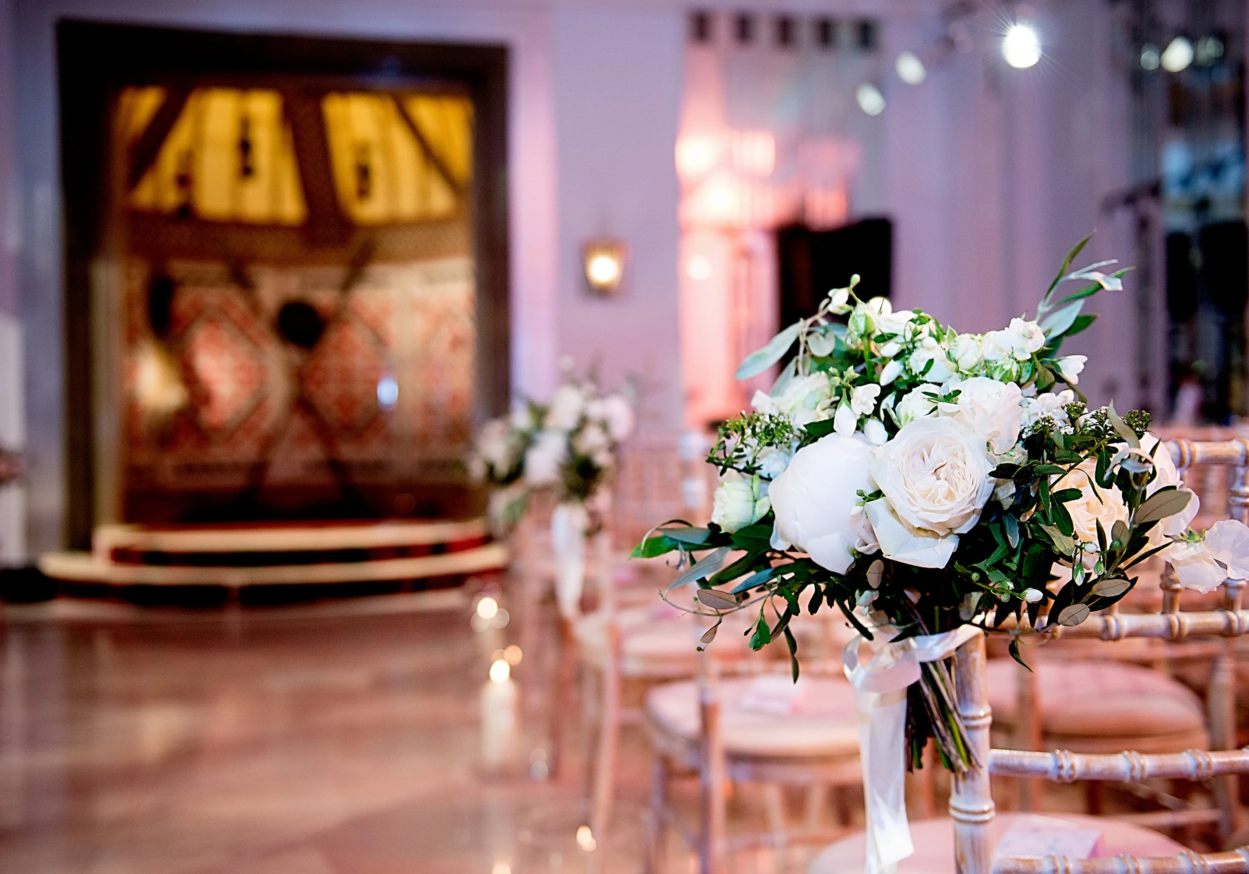 A floral centerpiece with white roses and greenery on a wooden chair at an indoor event with soft lighting.