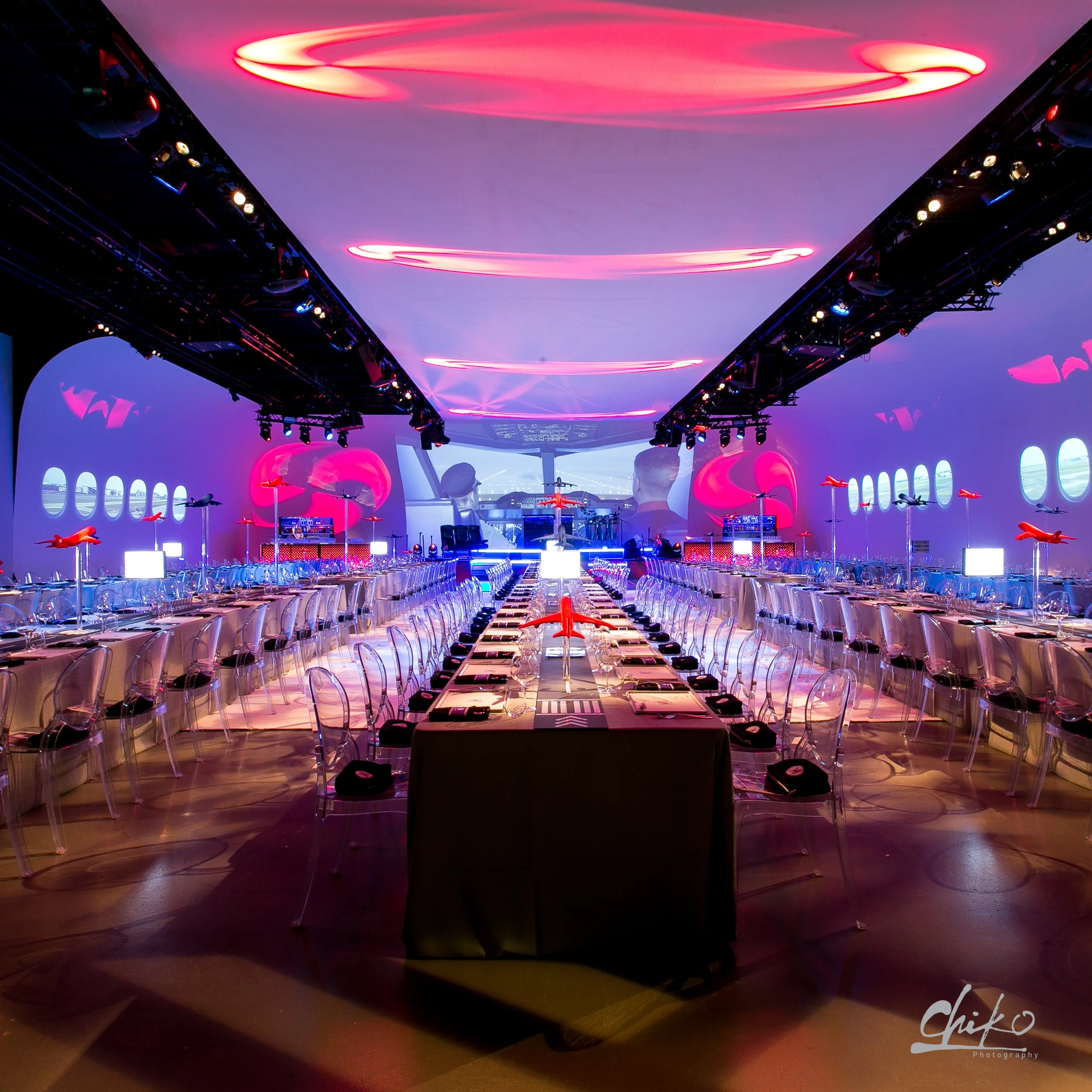 A dimly lit conference or event room with a long table set with chairs and red and pink ambient lighting overhead and along the sides.