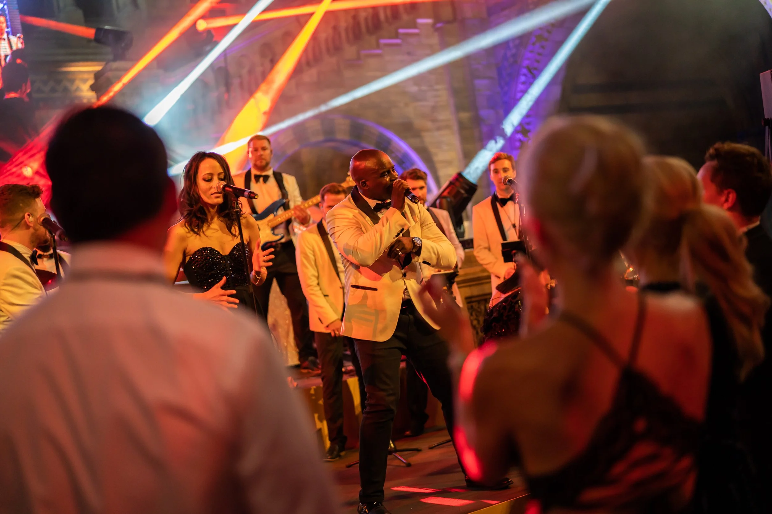 A lively stage performance featuring a man in a white tuxedo jacket singing into a microphone, surrounded by musicians and backup singers dressed in formal attire, with colorful lighting and an audience in the foreground at The Natural History Museum