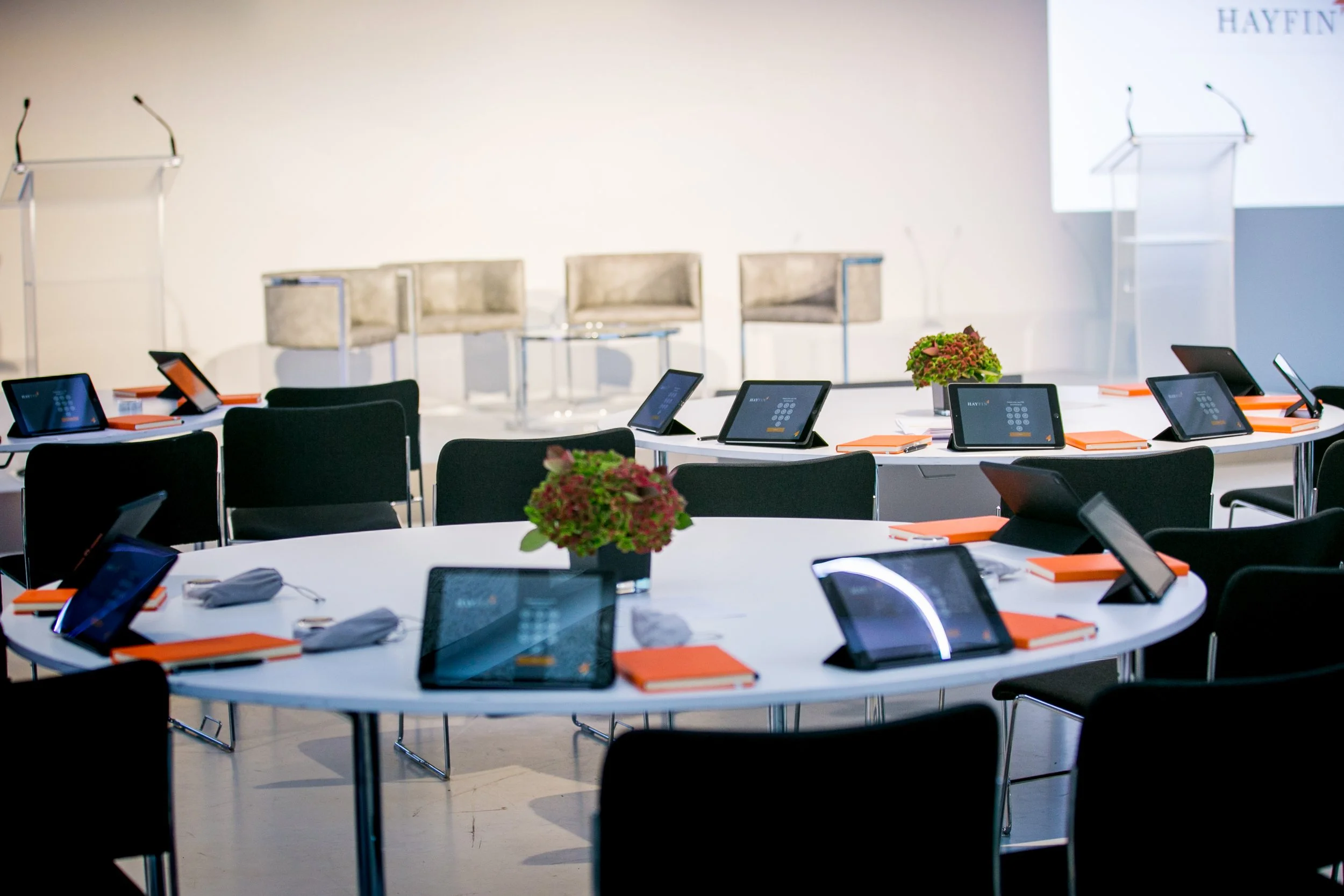 A conference room with round tables set up with tablets and orange notebooks, with chairs around each table. In the background, there are three beige chairs on a stage and a large screen with the text 'HAYFIN'.
