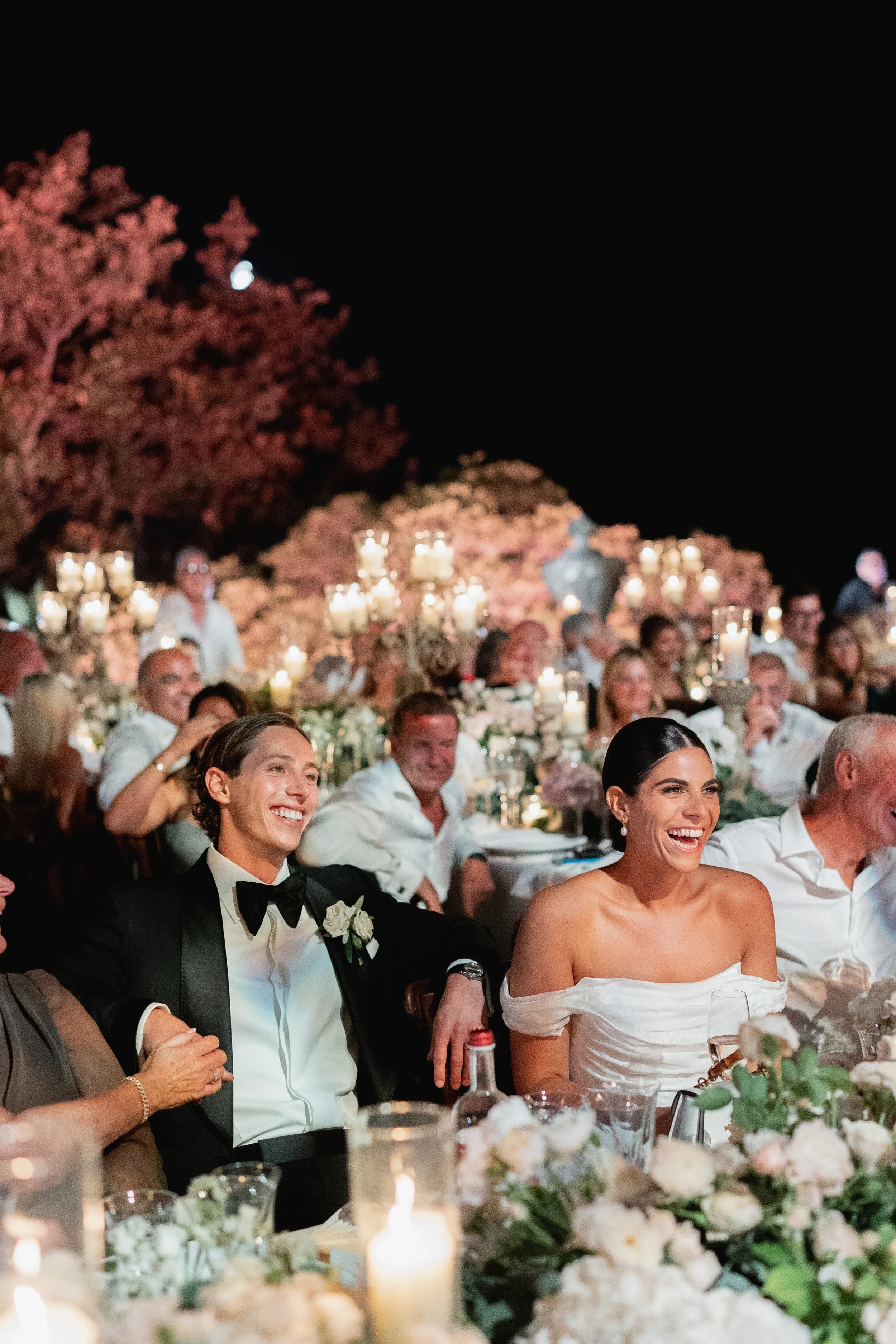 People seated at a wedding reception table smiling, with floral decorations and lit candles in the foreground, and a night sky with a visible moon in the background at Villa Palmieri in Florence