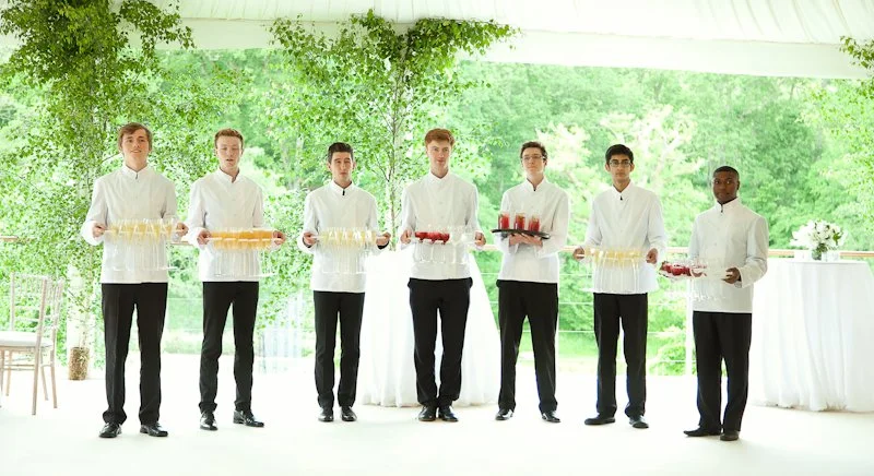 Seven waiters in white shirts and black pants standing in a row holding trays with drinks at a catered event.