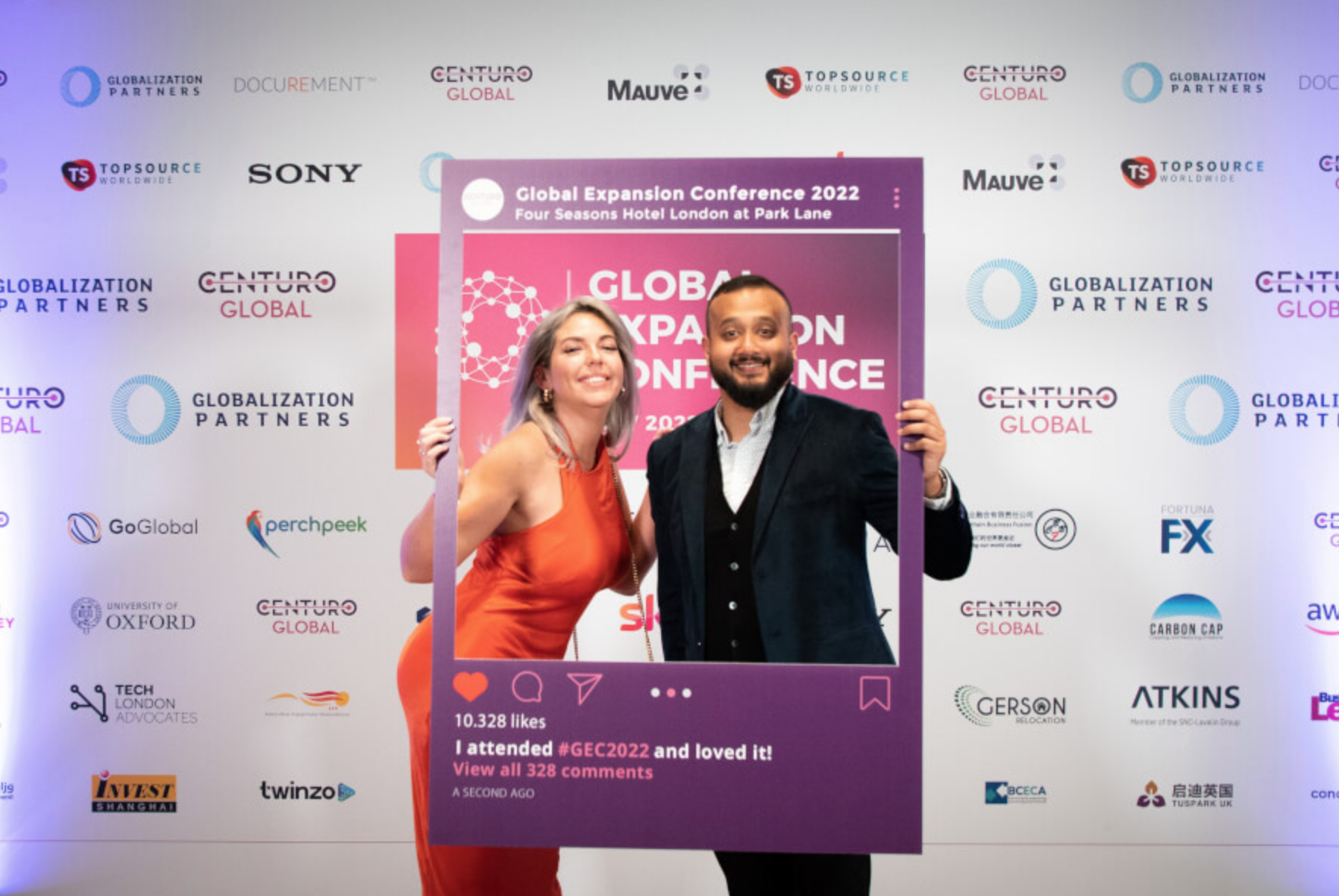 Two smiling people, a woman in an orange dress and a man in a dark suit, holding a purple frame with social media icons at a conference backdrop.