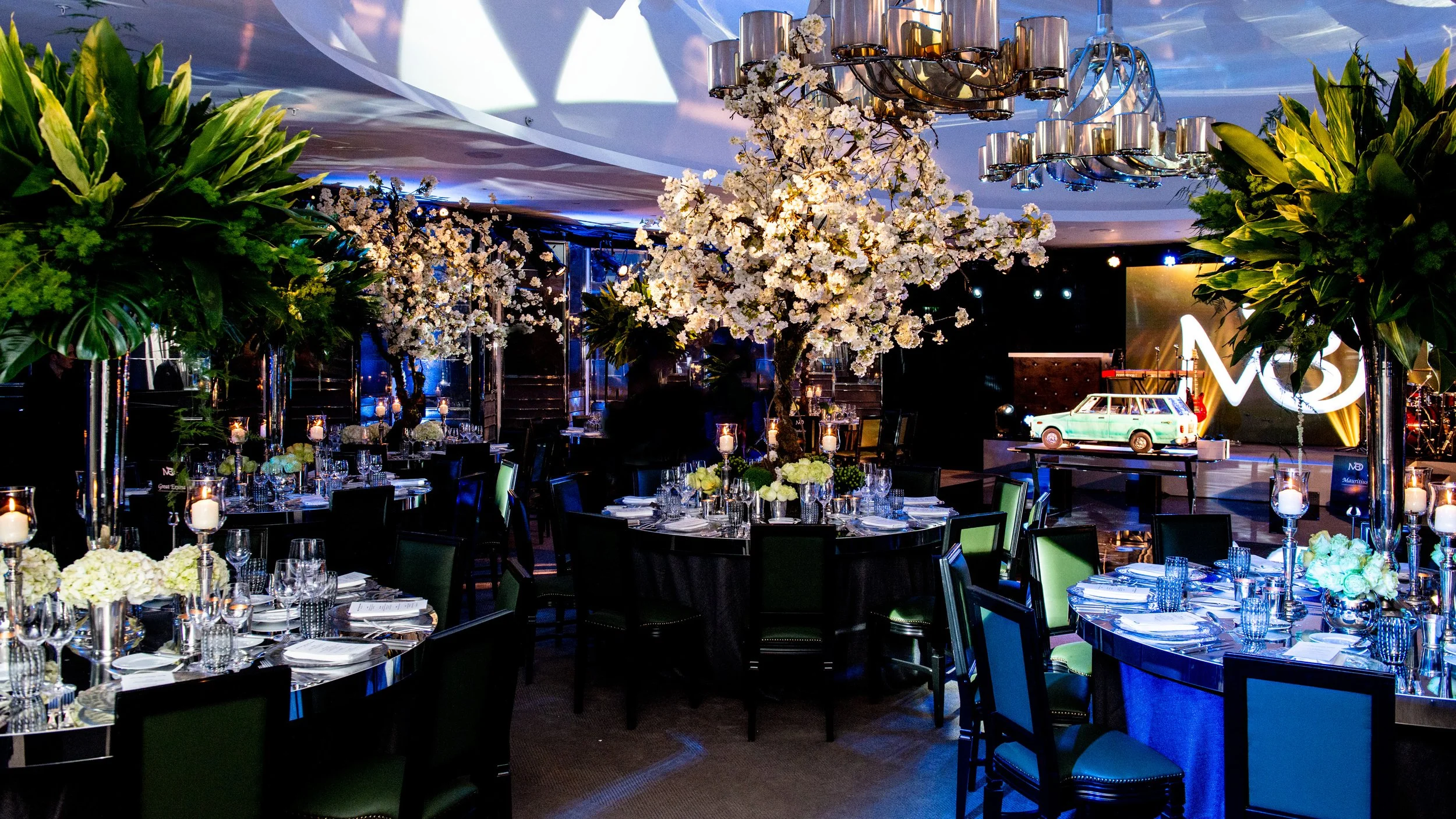 Luxury event space with floral centerpieces, decorated tables, and a stage with a vintage car display, under modern chandeliers and vibrant lighting.