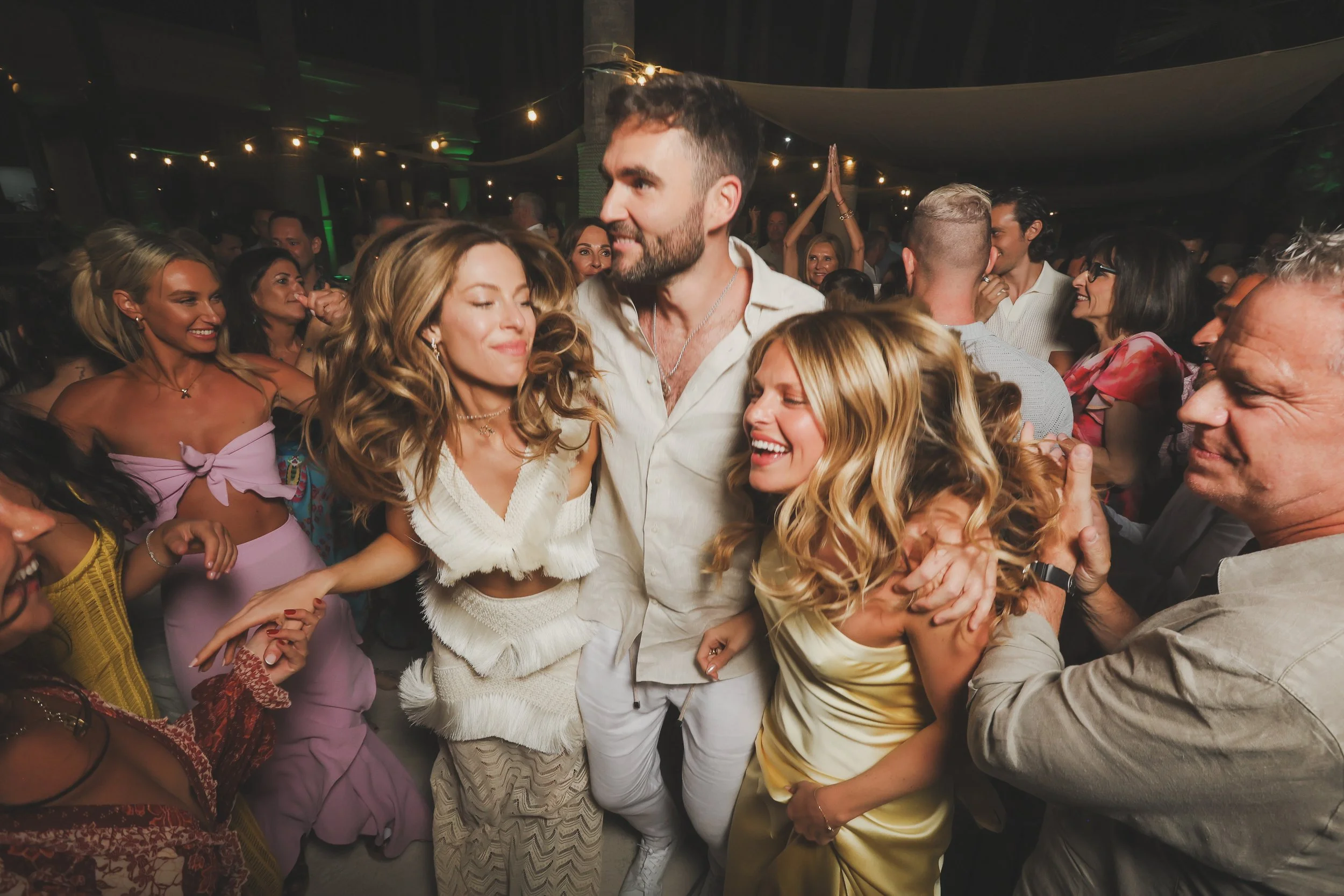 Group of people dancing and smiling at a lively party or celebration in a dimly lit indoor setting.