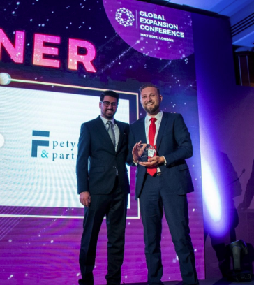 Two men in suits standing on a stage at the Global Expansion Conference, with one holding an award and the other smiling. Behind them is a large screen and conference branding.