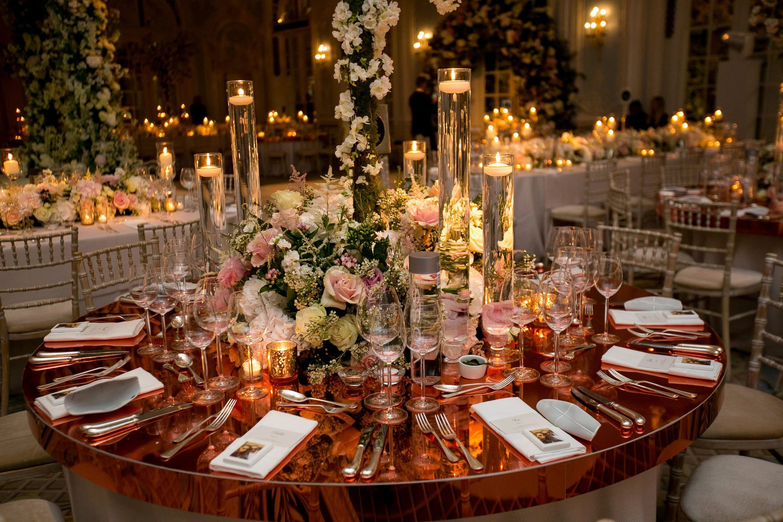 A round dining table decorated with elegant floral arrangements featuring pink and white flowers, tall candles in glass holders, smaller candles in gold holders, and neatly arranged place settings with white napkins and silverware. The background sho