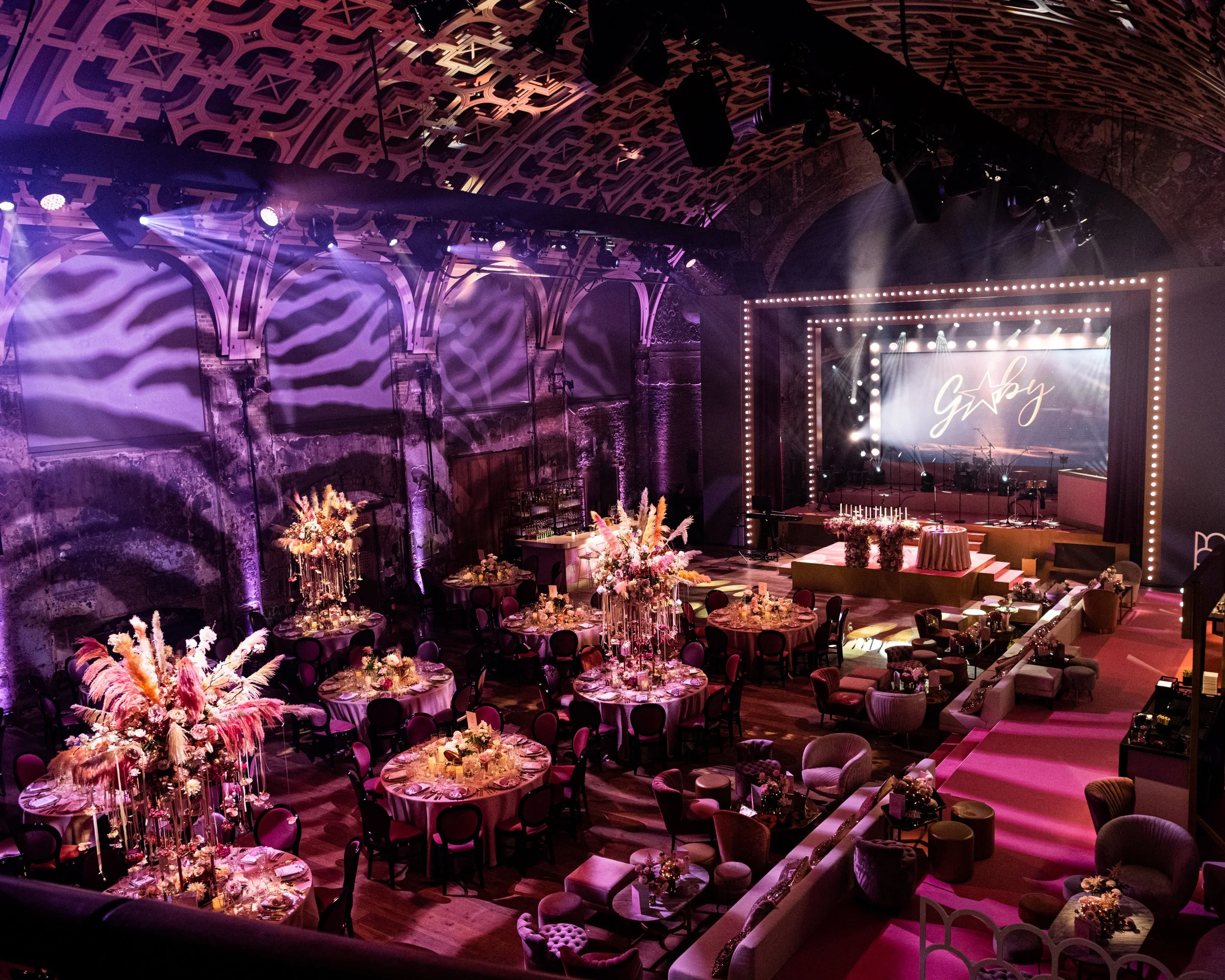 BAT MITZVAH AT BATTERSEA ARTS CENTRE The stage area has lighting effects, a screen with the word 'Gaby' and a star, and musical instruments. The ceiling features intricate geometric patterns, and the lighting creates a vibrant, festive atmosphere.