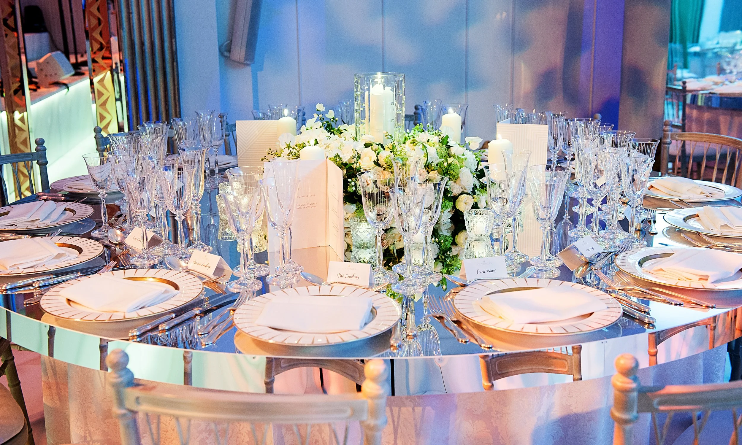 A round dining table decorated with white floral centerpiece, candles, wine glasses, and place settings with plates, napkins, and silverware, set for a formal event or celebration.