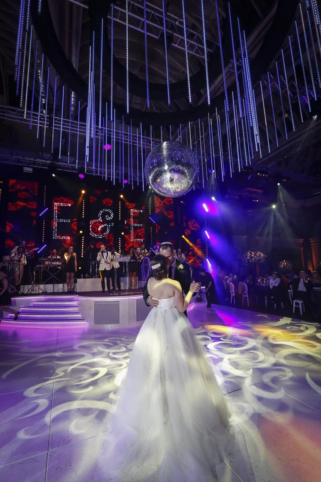 A bride and groom dance at their wedding reception under colorful lighting and a large disco ball, with a live band performing on stage in the background.