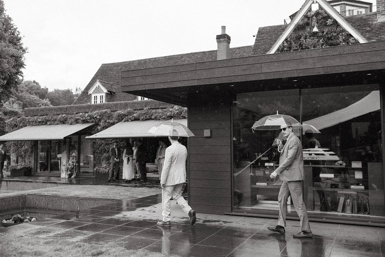 A black and white photo of a modern house with large glass windows and a wooden exterior, where several people are gathered outside on a rainy day. Some are standing under awnings, and one man is walking across the patio while another man is drinking