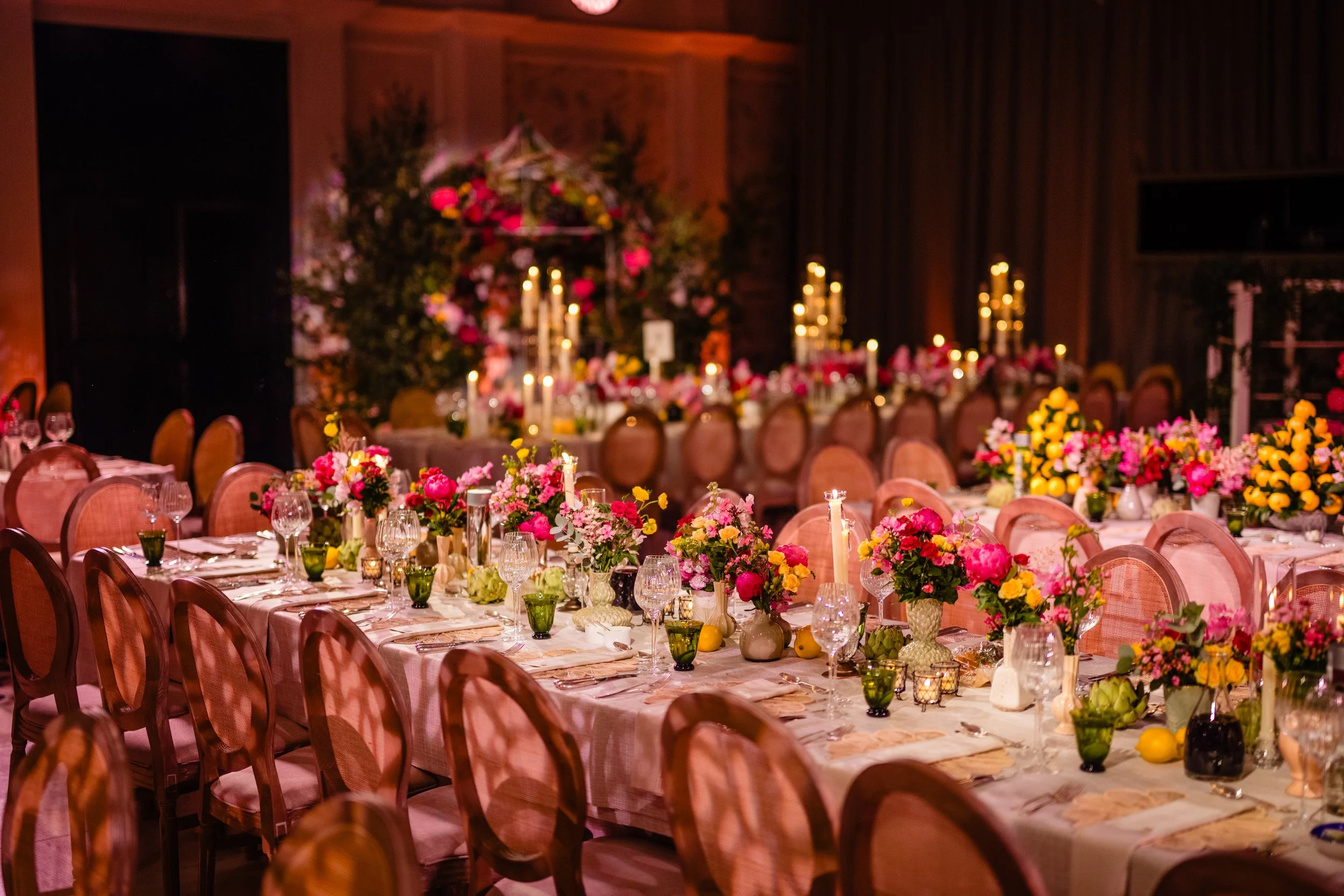 A banquet hall decorated with pink, yellow, and purple flowers, lit by candlelight, with framed floral art and dark wood paneling on the walls.