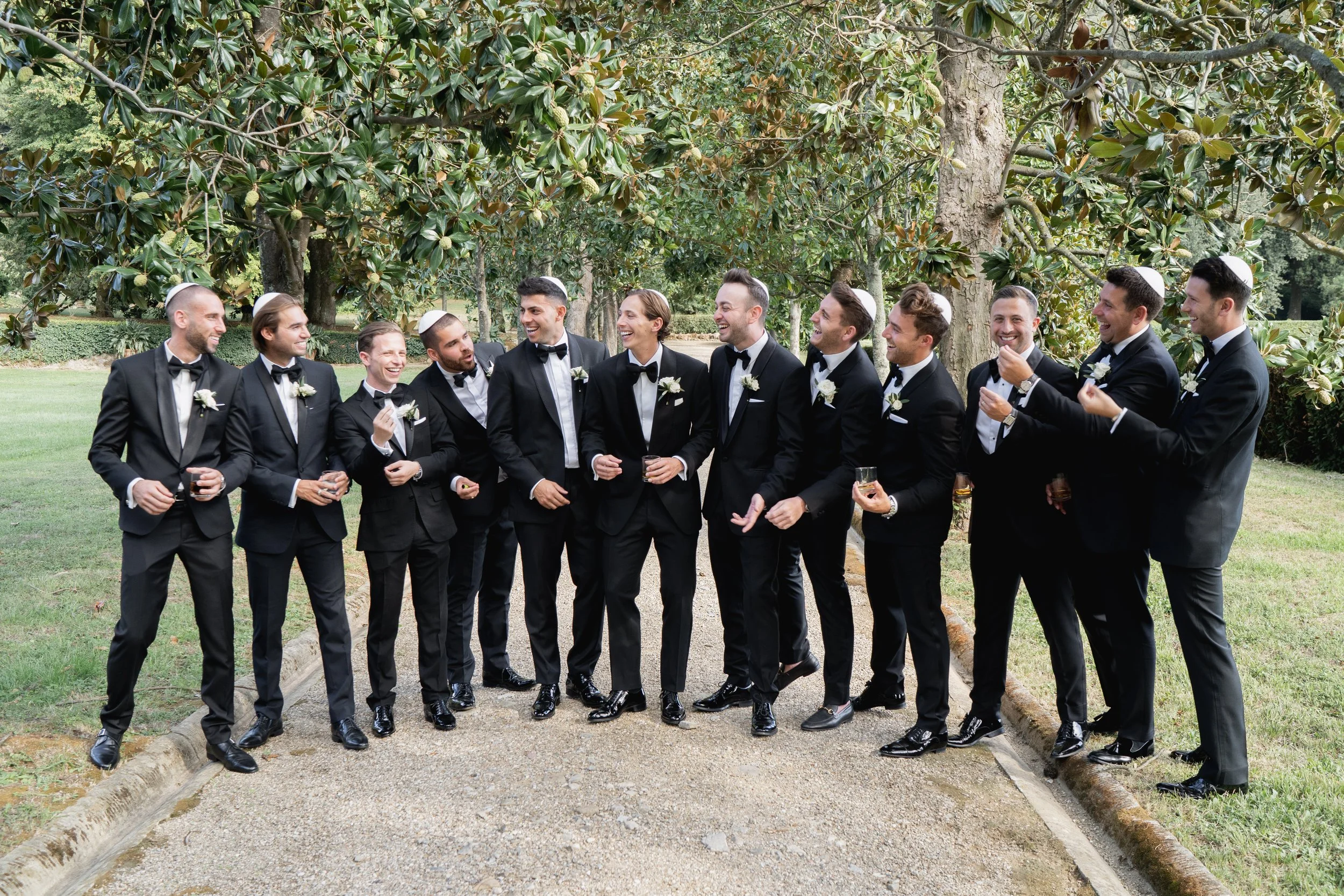 Group of men dressed in black tuxedos with white shirts and black bow ties standing outdoors, laughing and engaging with each other, some wearing white kippahs, with lush green trees in the background.