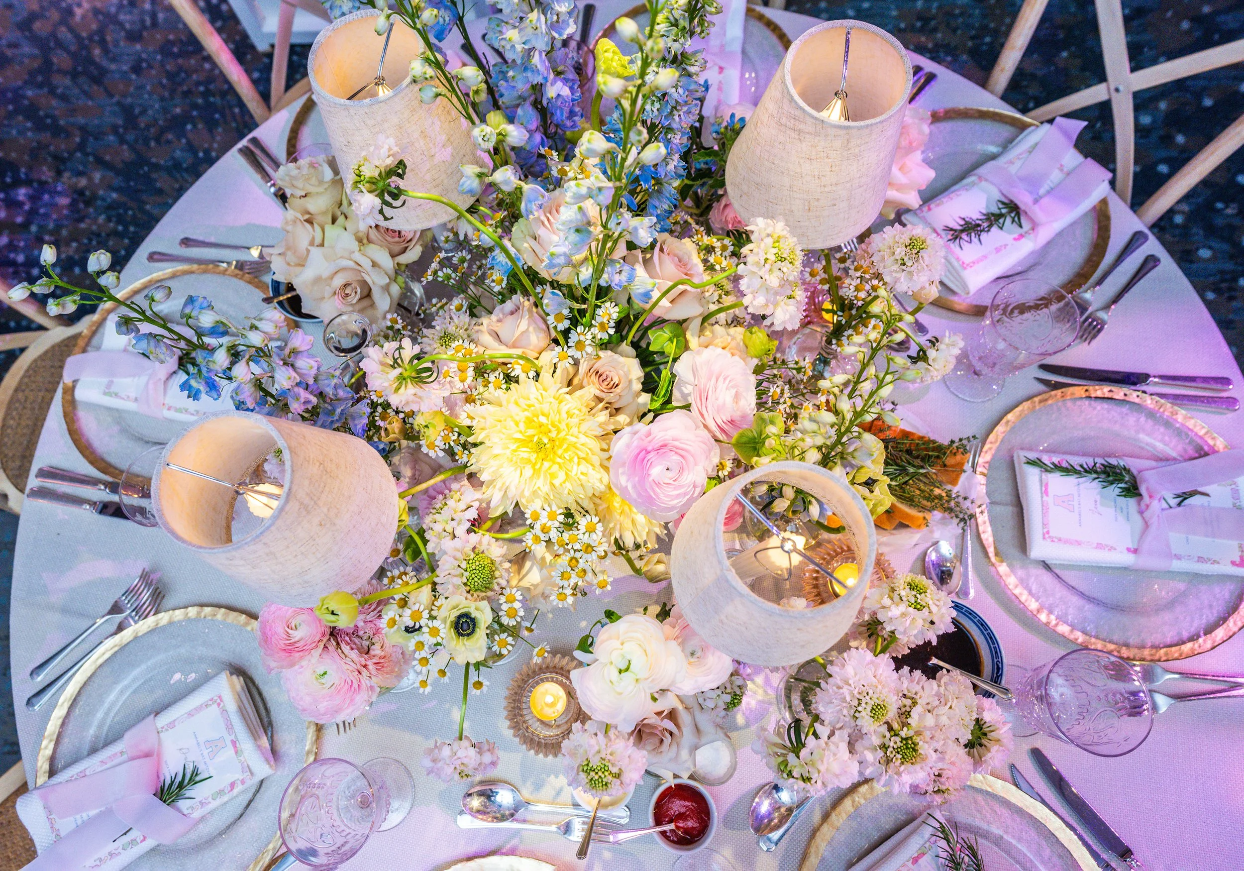 Overhead view of a floral table centerpiece with pink, white, and purple flowers, three white lamps, place settings with pink napkins, glasses, and silverware, small candles, and a pink menu or place card on a white tableclot.