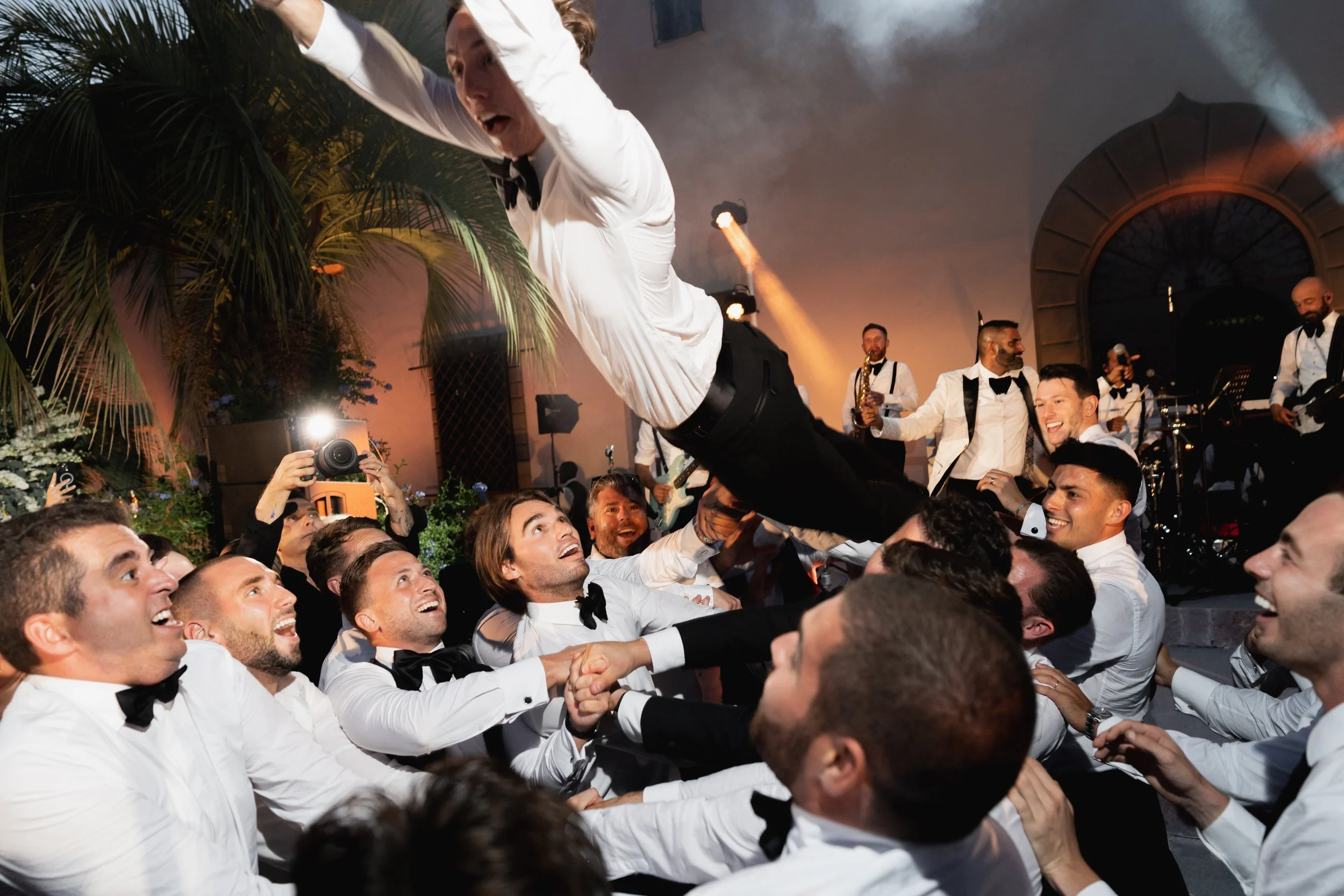A group of men dressed in white shirts and black bow ties are lifting a man dressed in a tuxedo  The man being lifted appears excited or surprised. A band playing music and tropical plants are visible at Villa Palmieri in Florence.