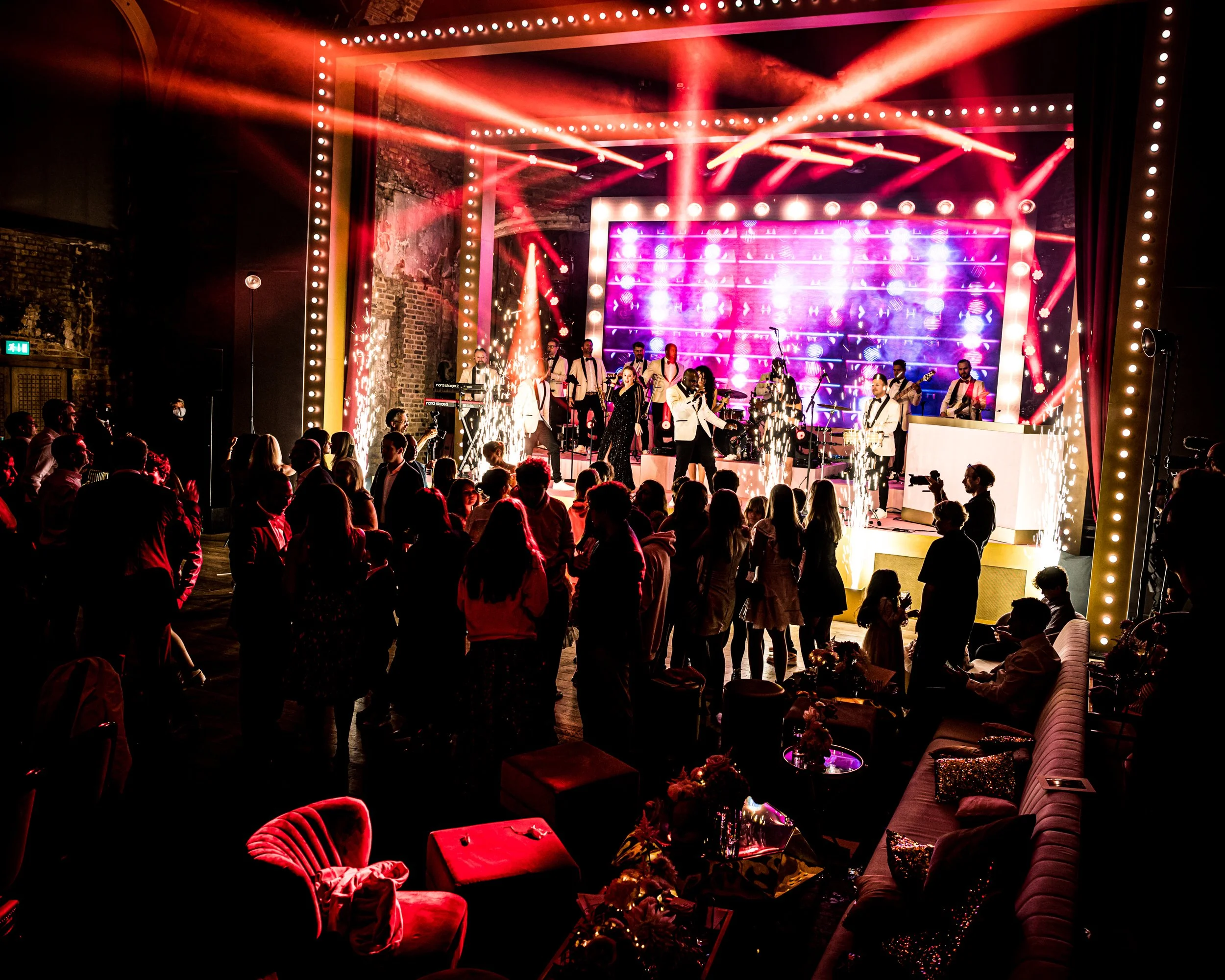 A lively indoor concert or event with a stage illuminated by colorful lights, featuring musicians and a singer, with an audience watching from tables and chairs. BAT MITZVAH AT BATTERSEA ARTS CENTRE