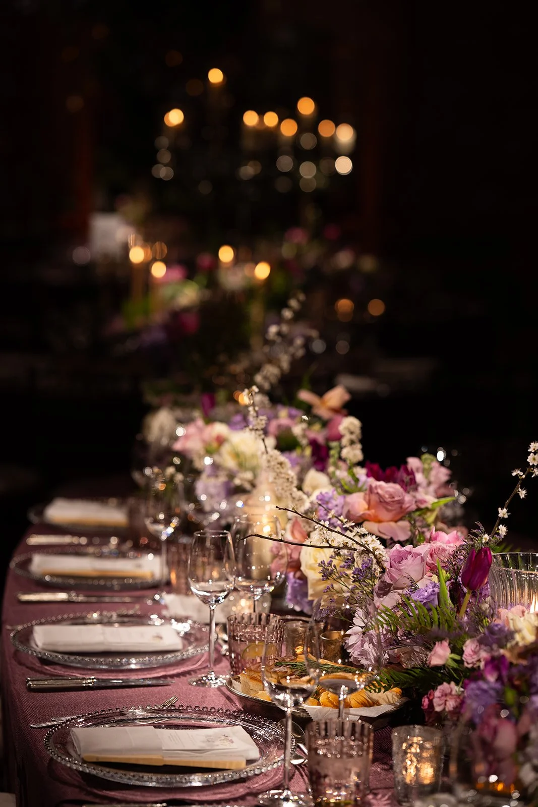 Elegant dining table decorated with pink, purple, and white flowers, glassware, and candles in a dimly lit setting with blurred candles in the background at The Peninsula London